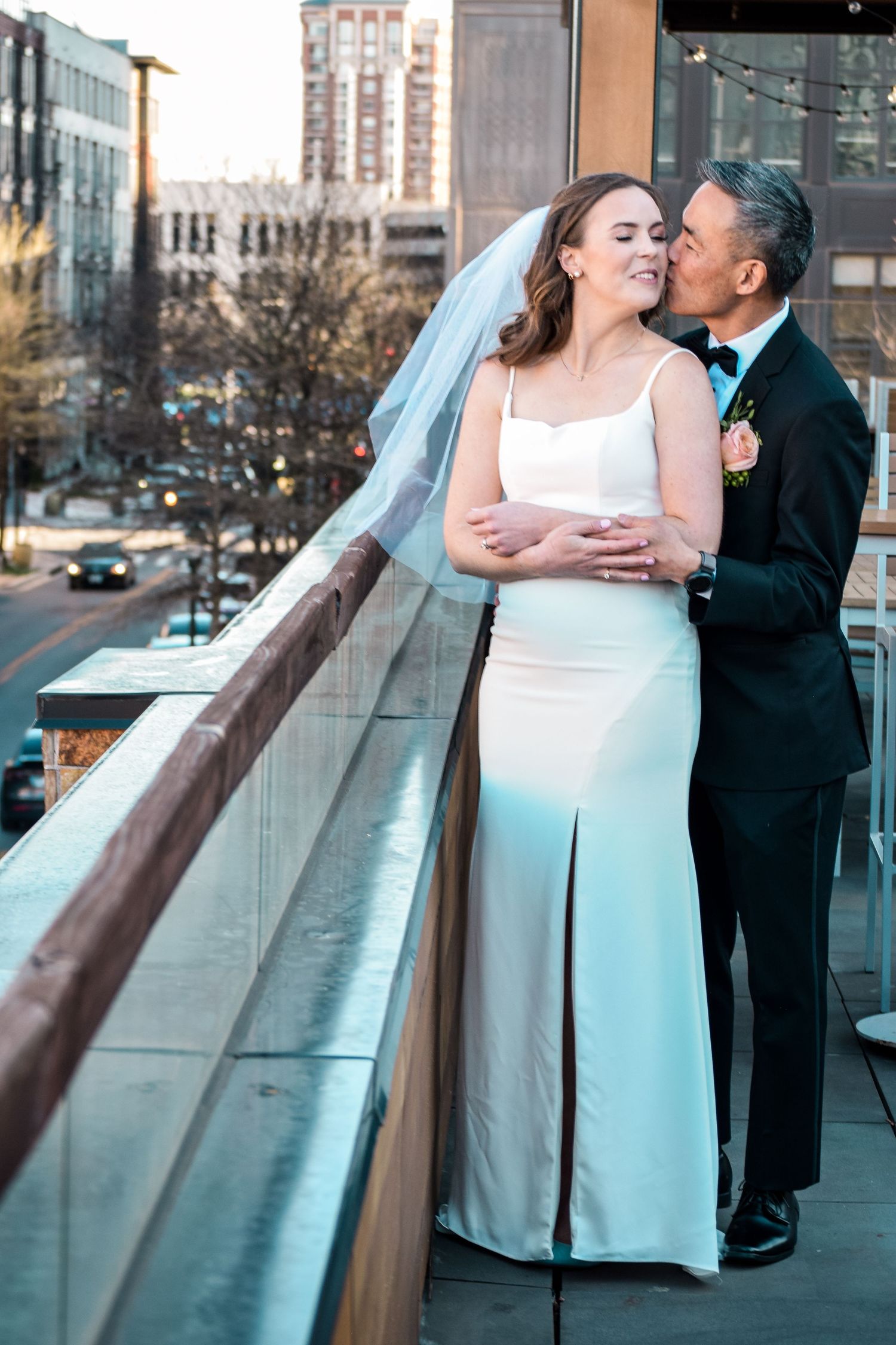 Groom tenderly kisses smiling bride on cheek while embracing her on rooftop wedding venue.