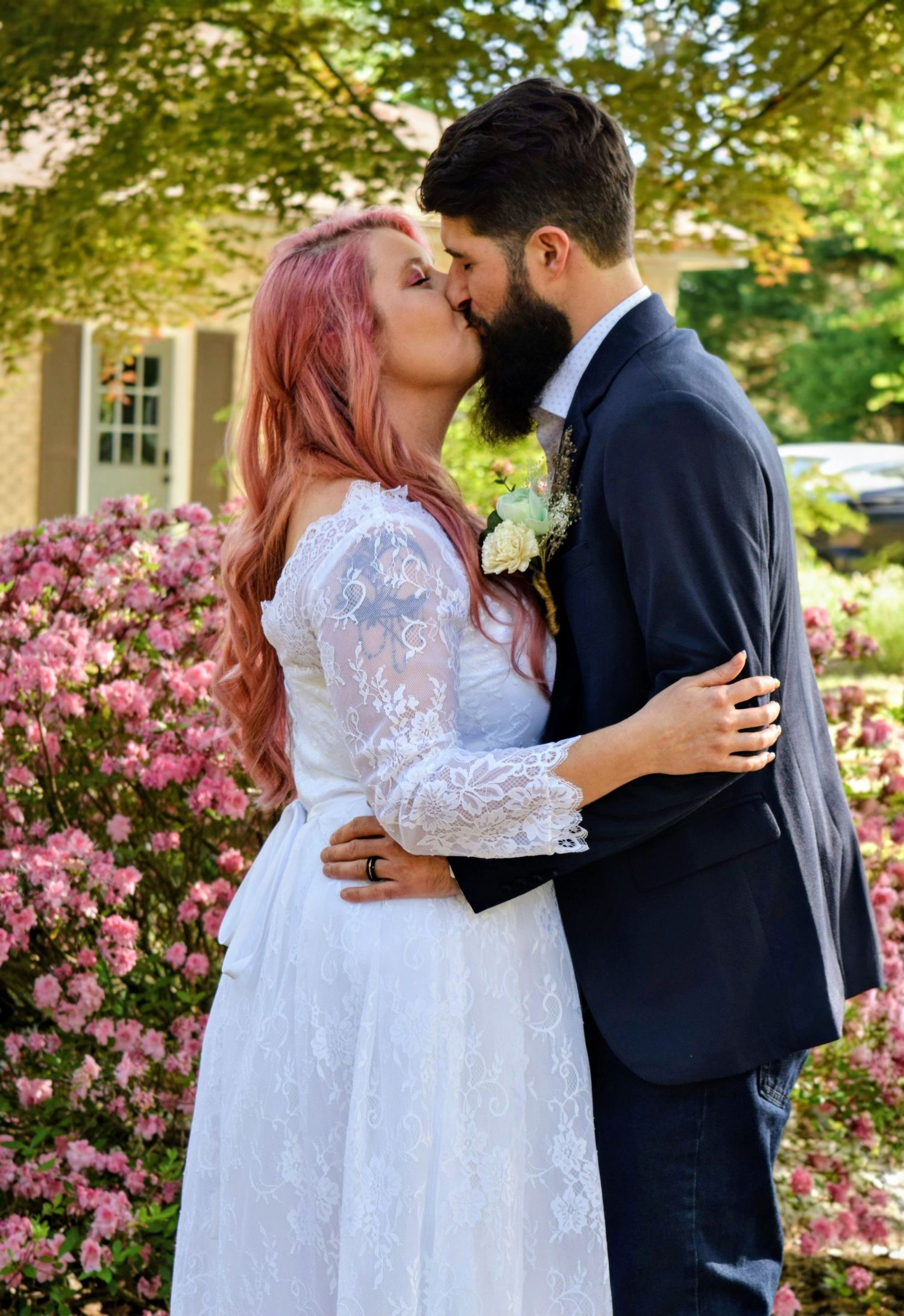 Bride with pink hair and groom with beard sharing a kiss among pink flowers, bride wearing lace wedding dress.