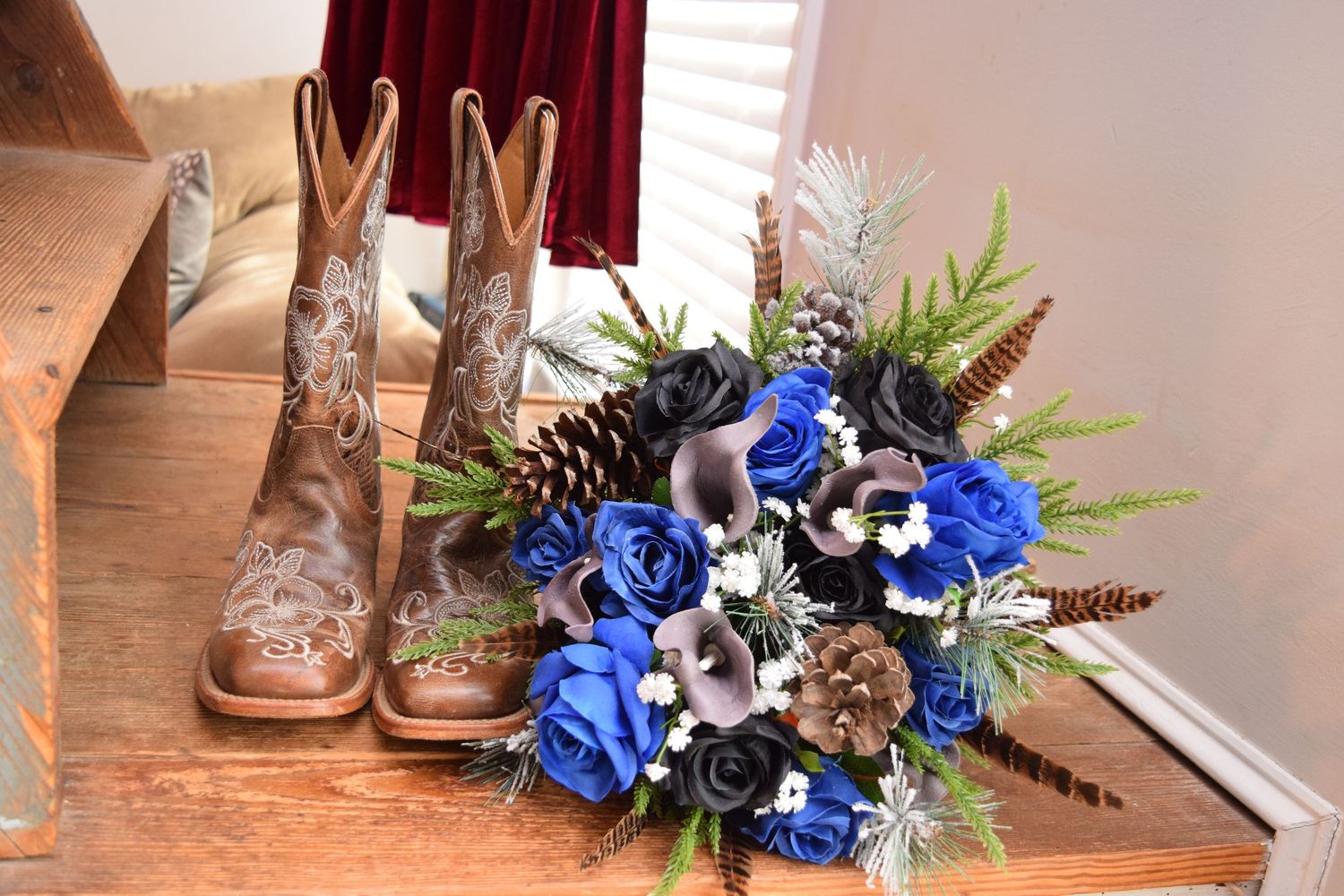 Blue and black bridal bouquet with roses and pinecones displayed next to tan cowboy boots on wooden stairs.