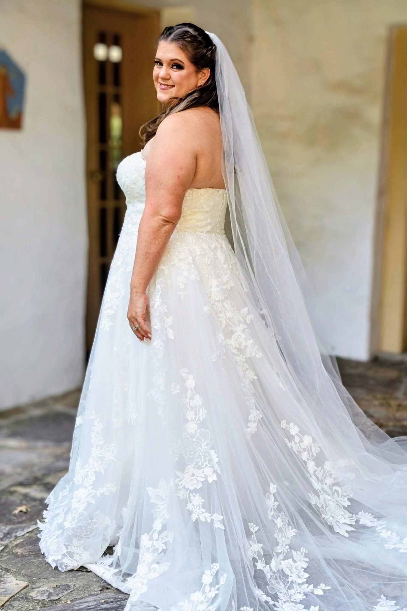 Elegant bride in strapless lace wedding gown with long veil and floral train posing outdoors.