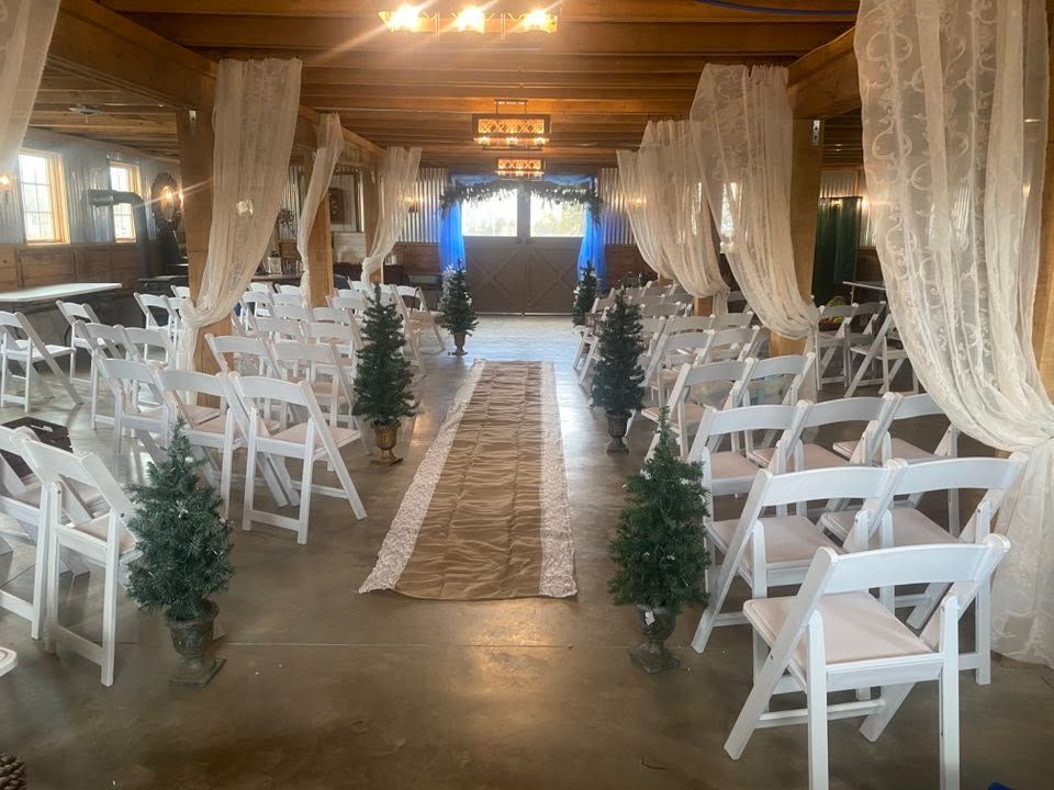 Elegant barn wedding ceremony aisle with white chairs, burlap runner, and small Christmas trees.