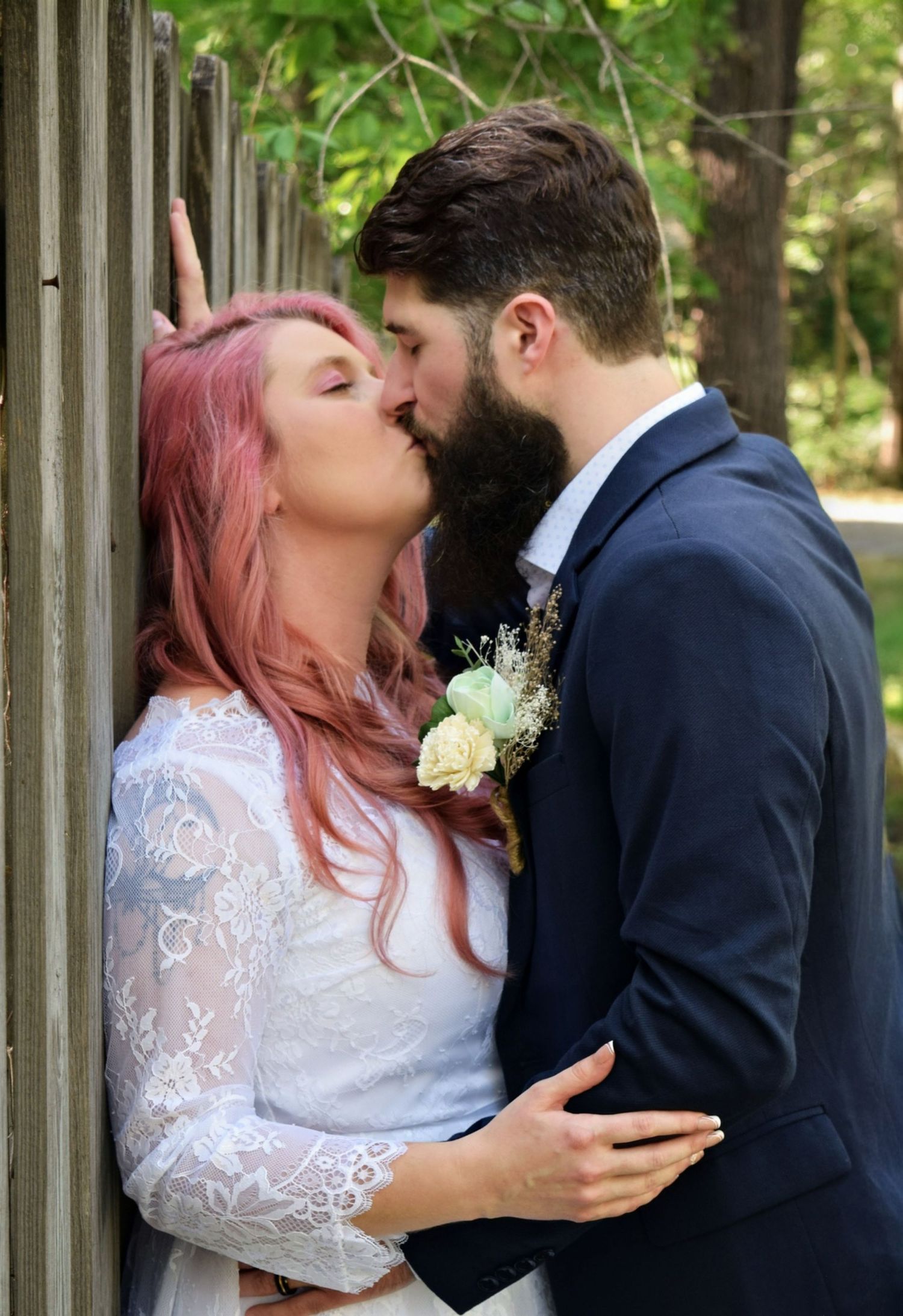 Bride with pink hair and bearded groom sharing a romantic kiss against a wooden fence, both in wedding attire.