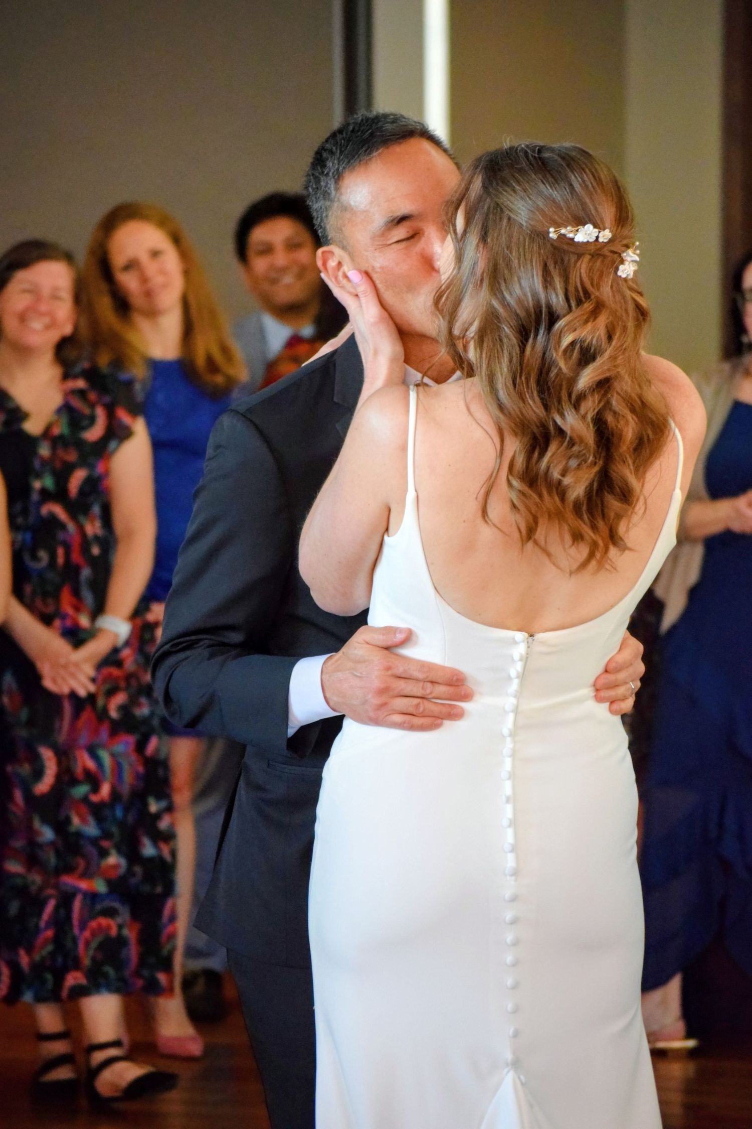 Groom kisses bride during first dance while wedding guests celebrate in background.
