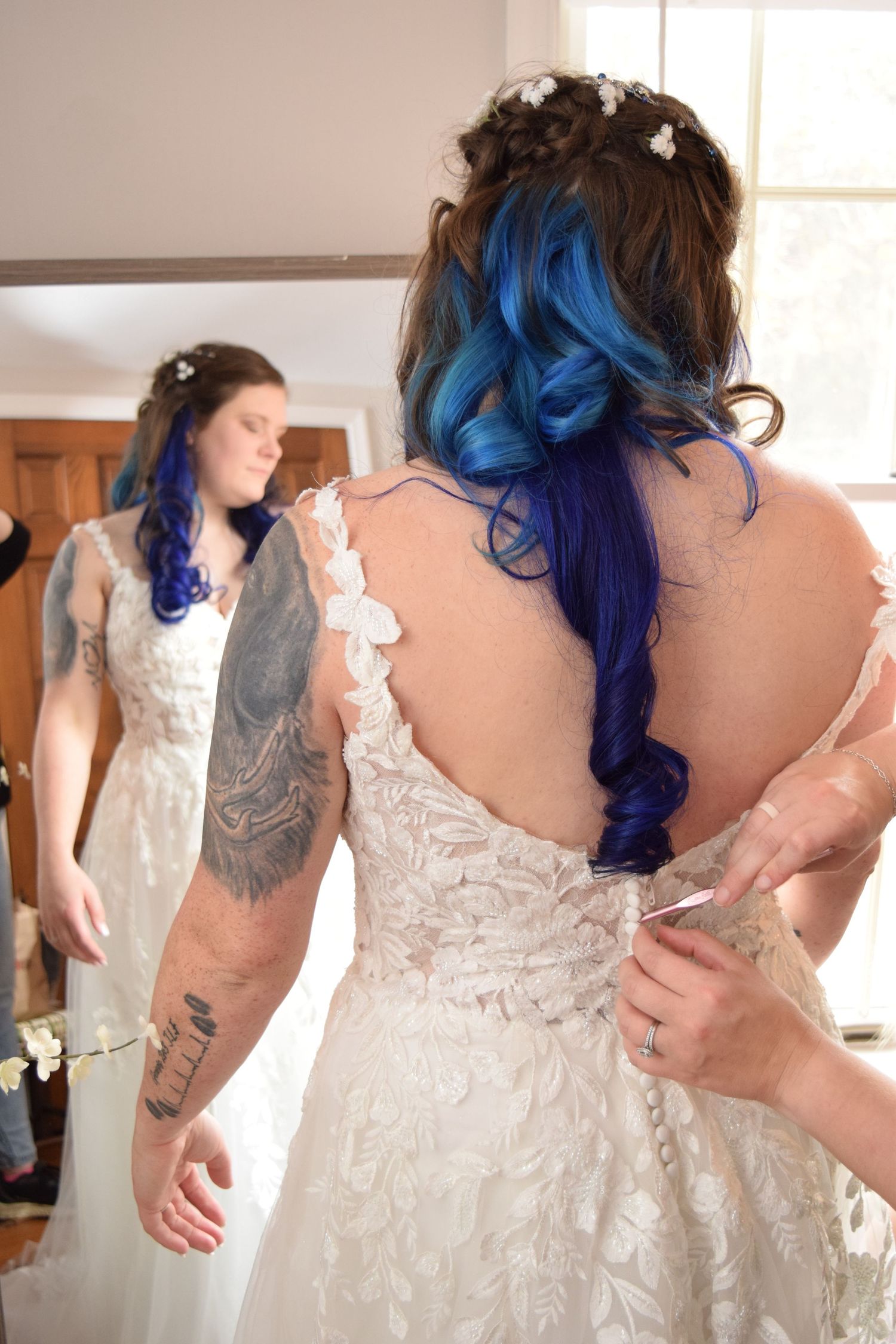 Bride with blue ombre hair and tattoos getting wedding dress buttoned up from behind.