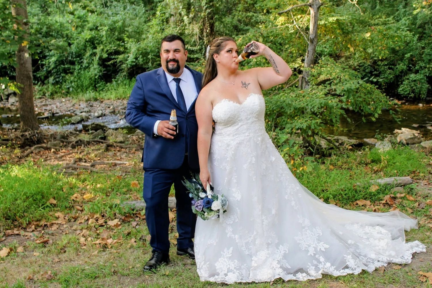 Bride and groom celebrating outdoors near creek, bride drinking champagne in lace gown.