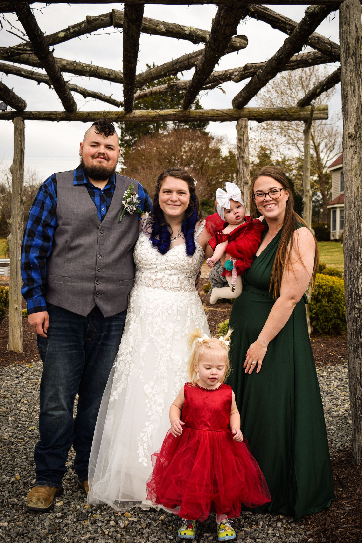 Wedding couple posing under rustic wooden arbor with bridesmaid in green dress holding baby in red.