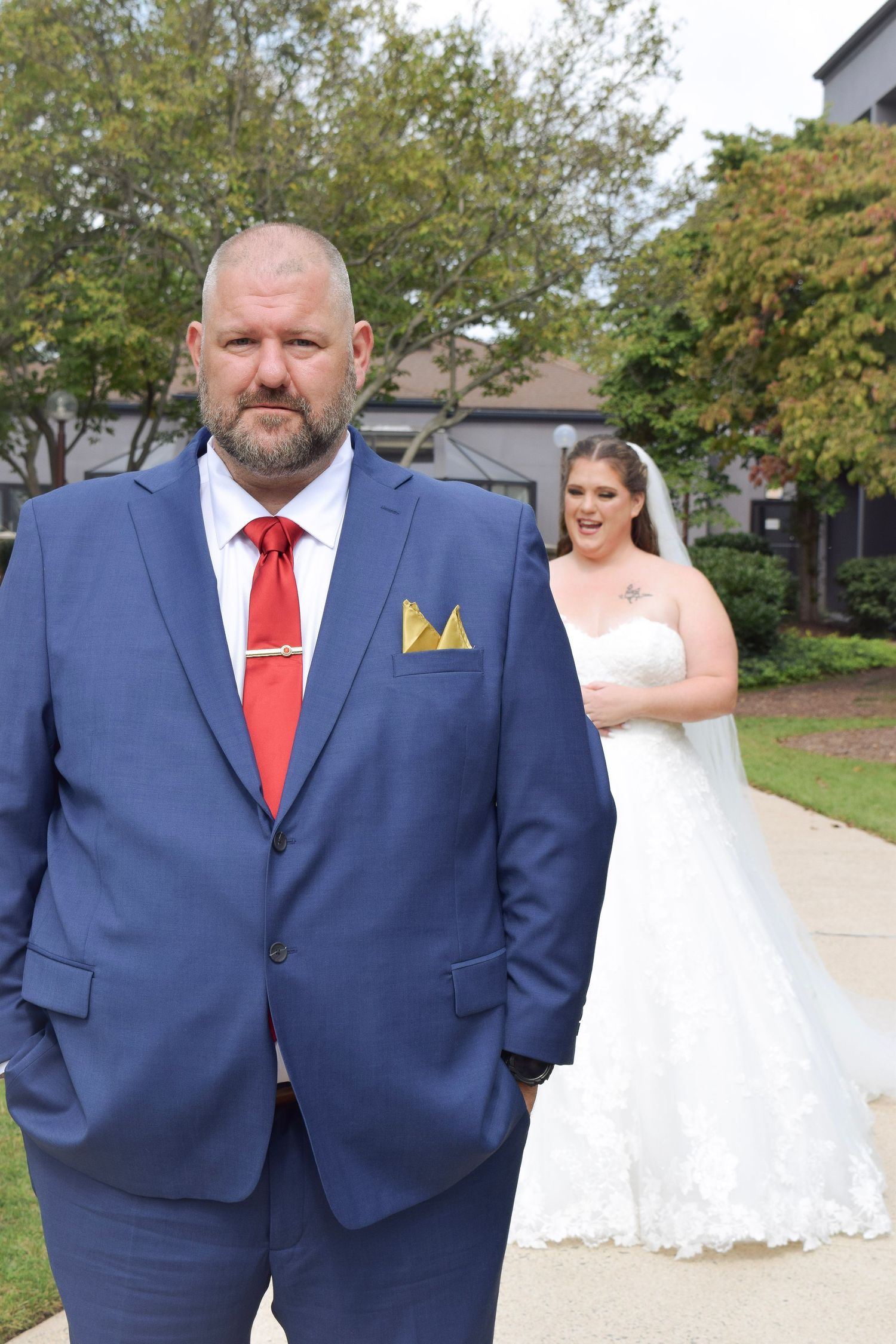 Groom in navy blue suit with red tie standing outdoors as bride in white wedding gown approaches behind him.