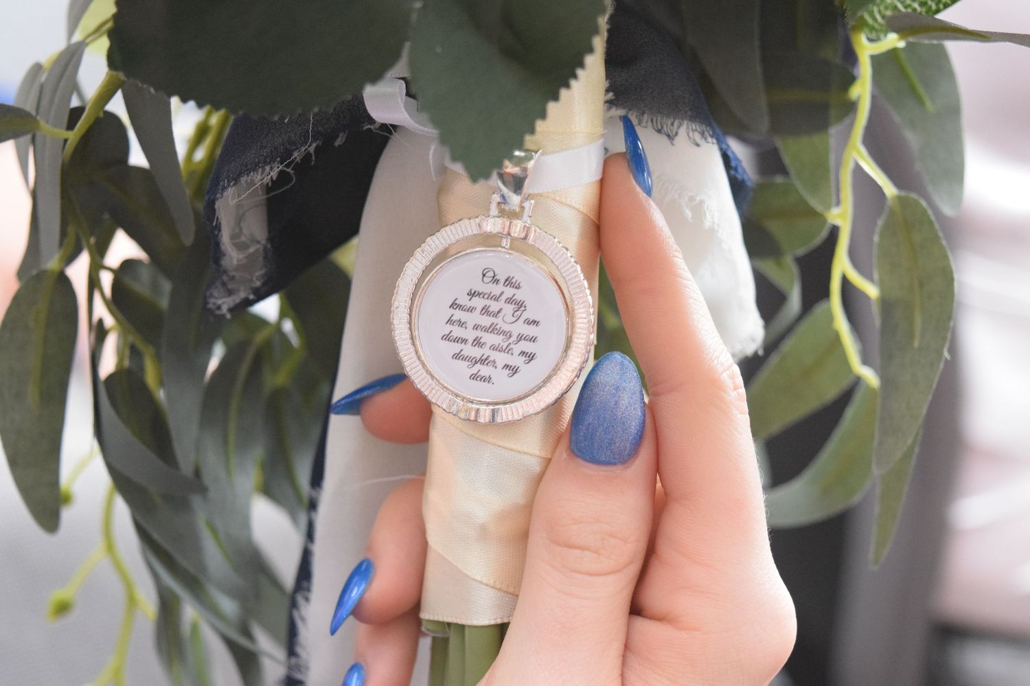 Close-up of a hand holding a round locket pendant with handwritten text, surrounded by wedding bouquet greenery.