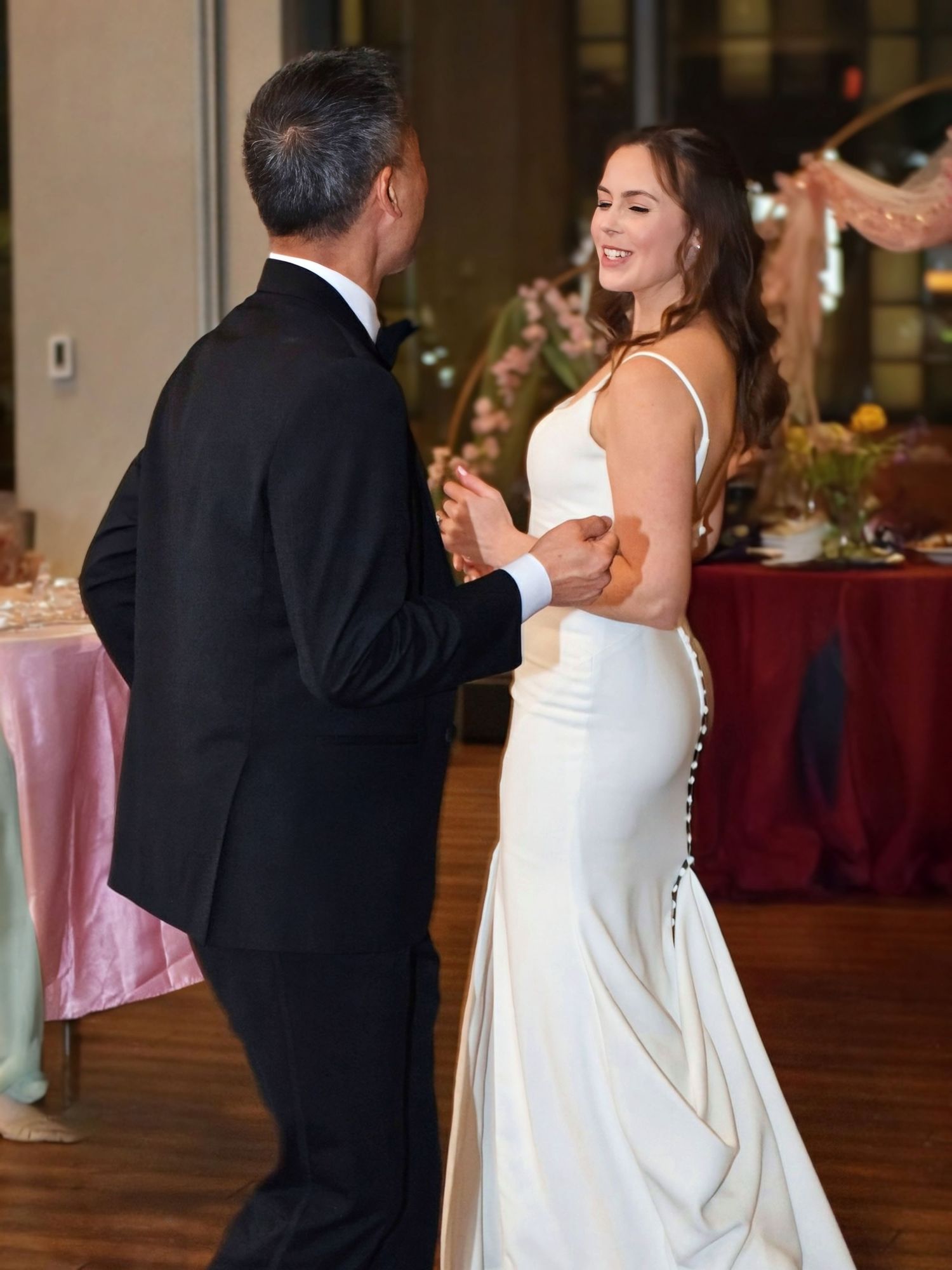 Joyful bride in a fitted white gown with button back dances with groom at wedding reception.