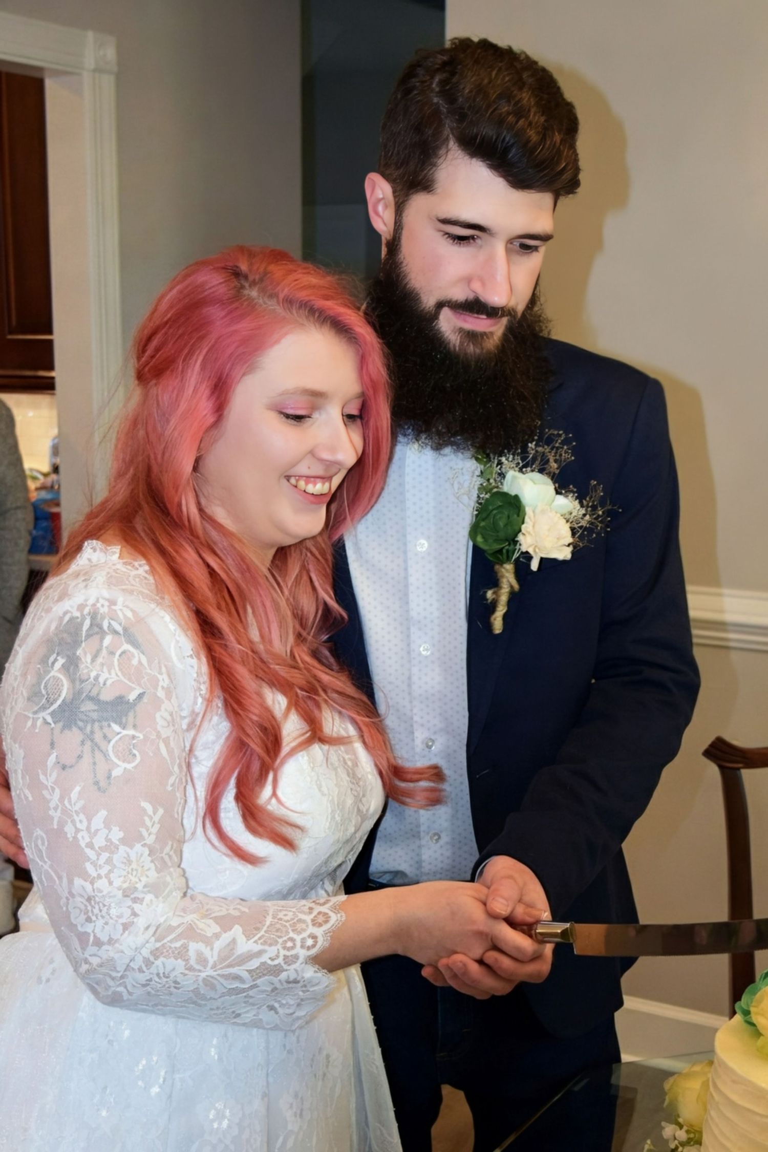 Smiling bride with pink hair and bearded groom cutting wedding cake together at indoor wedding reception.