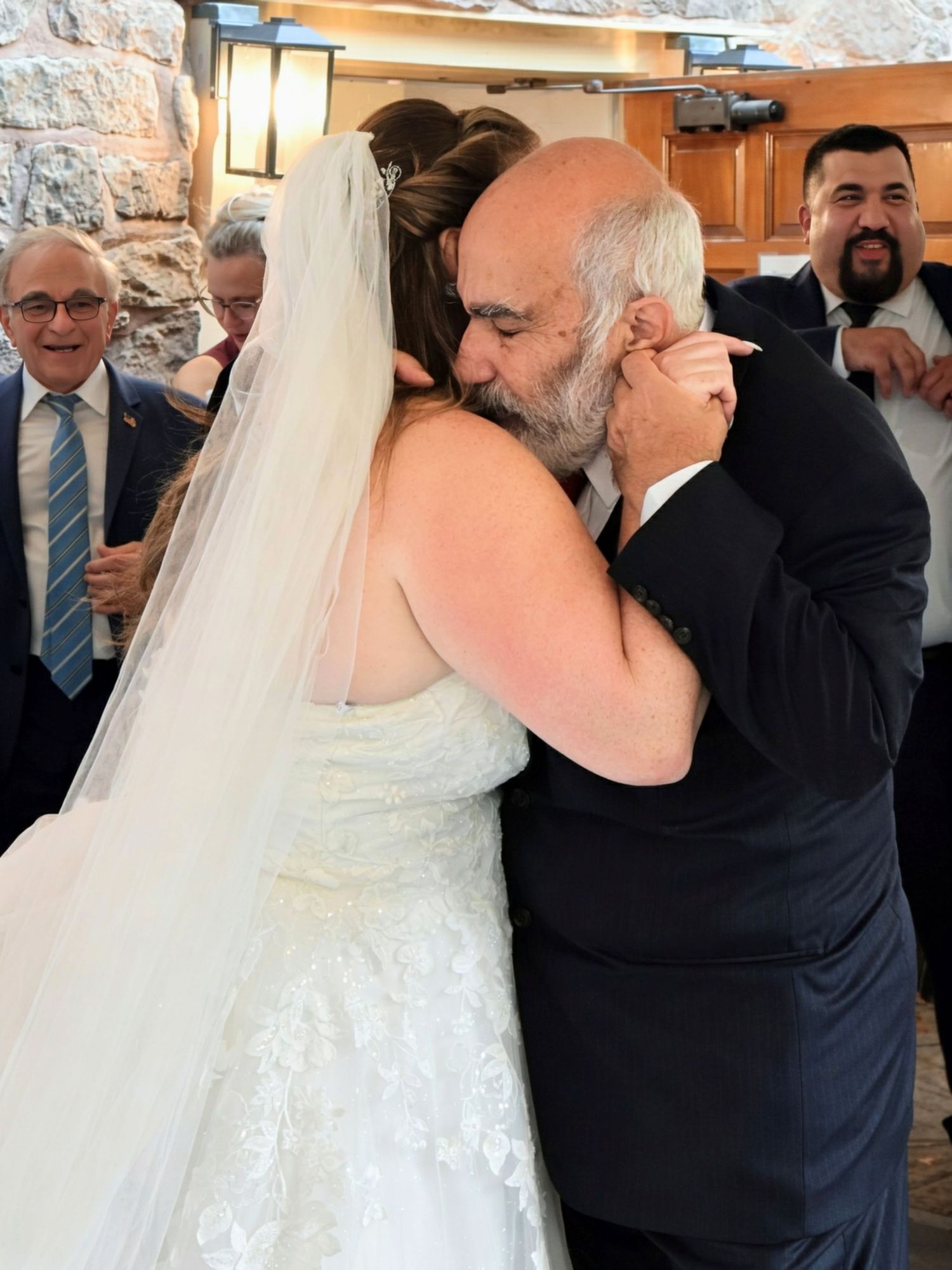 Bride in white lace wedding gown sharing tender kiss with older man at reception.