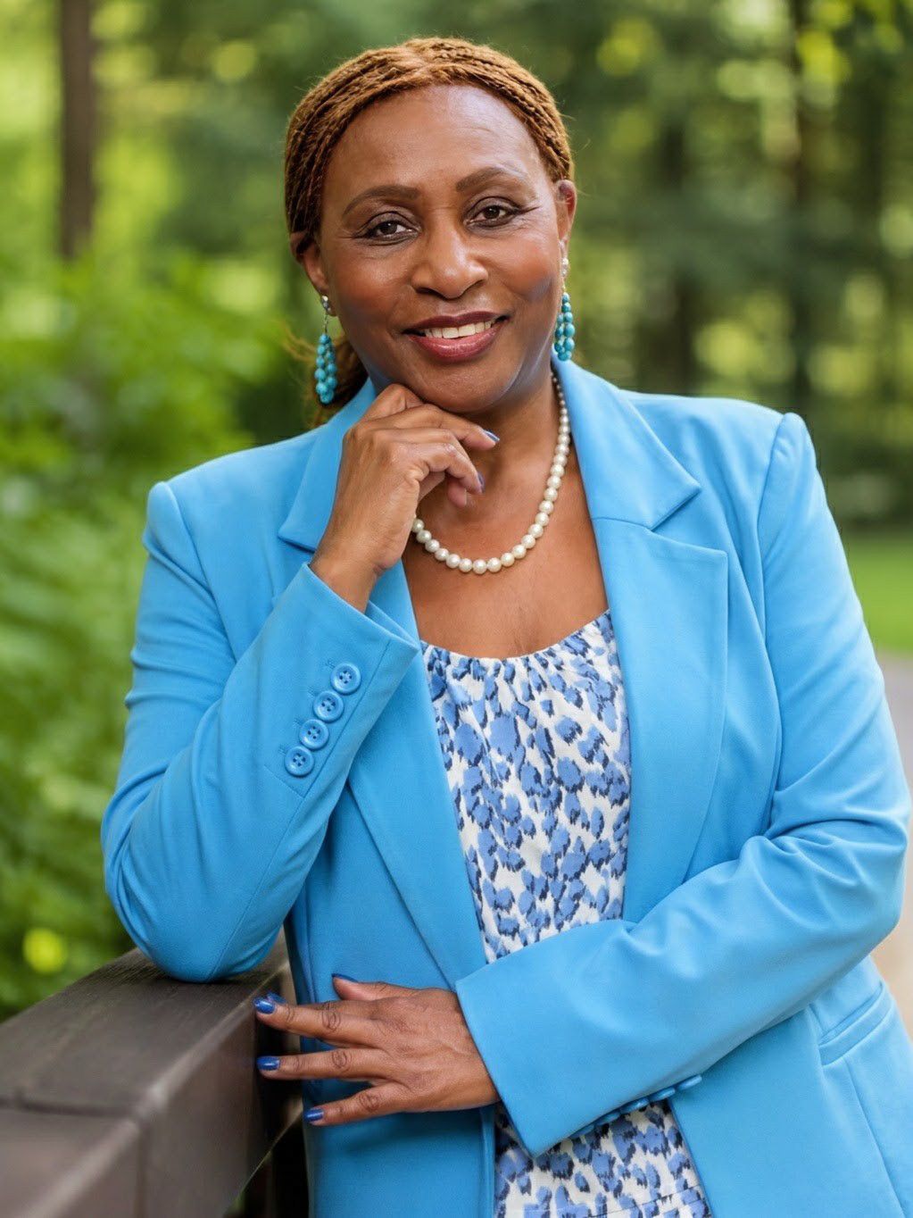 Professional woman in blue blazer and pearl necklace smiling outdoors with green trees in background.