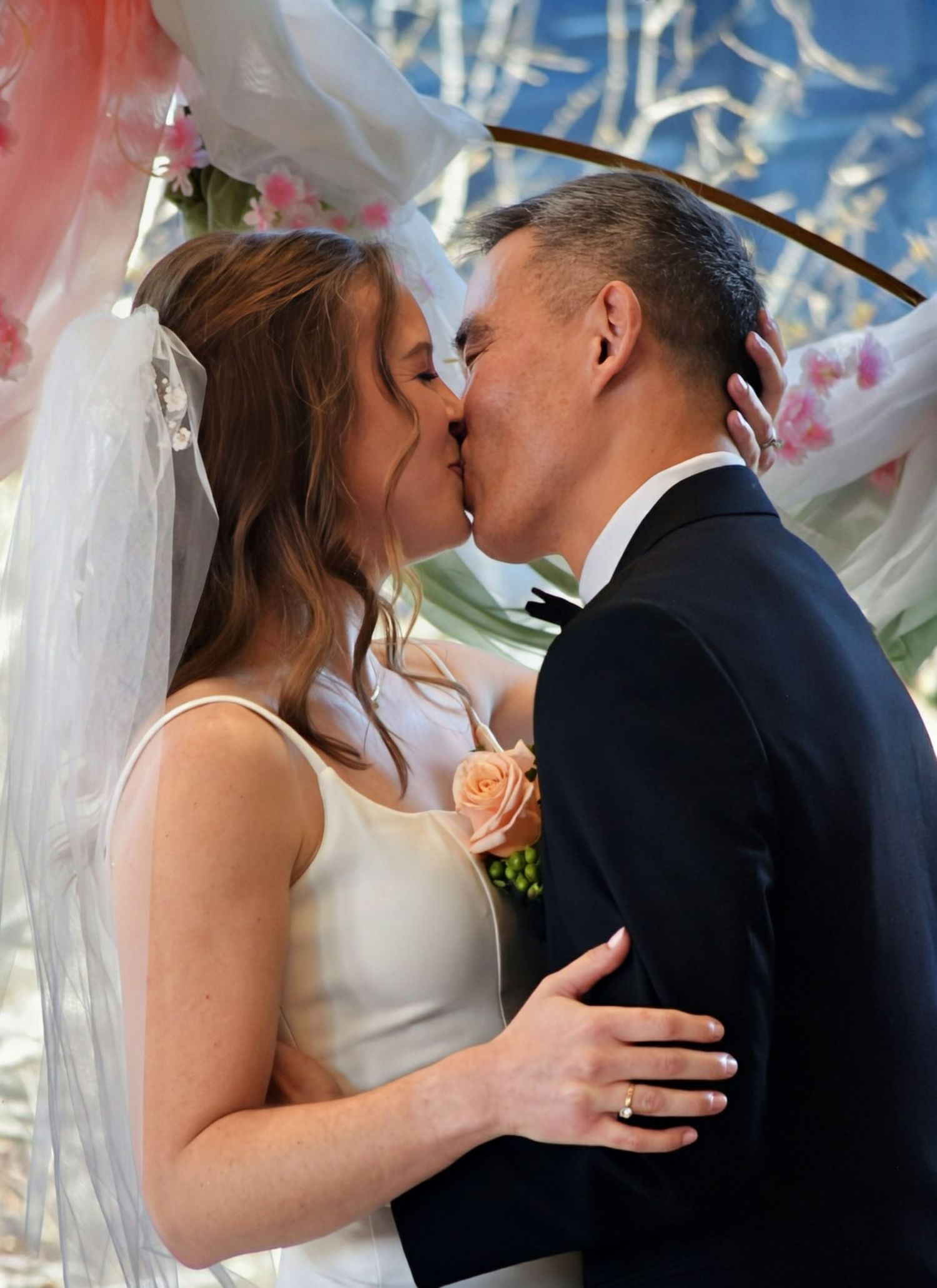 Bride and groom share first kiss under pink floral wedding arch during ceremony.
