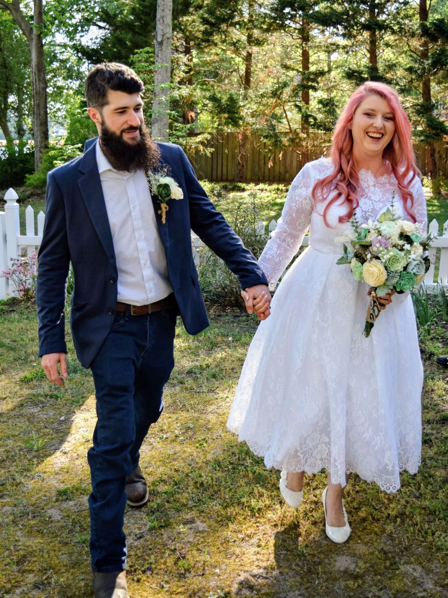Smiling newlywed couple holding hands outdoors, pink-haired bride in lace dress with bouquet and bearded groom in navy suit.