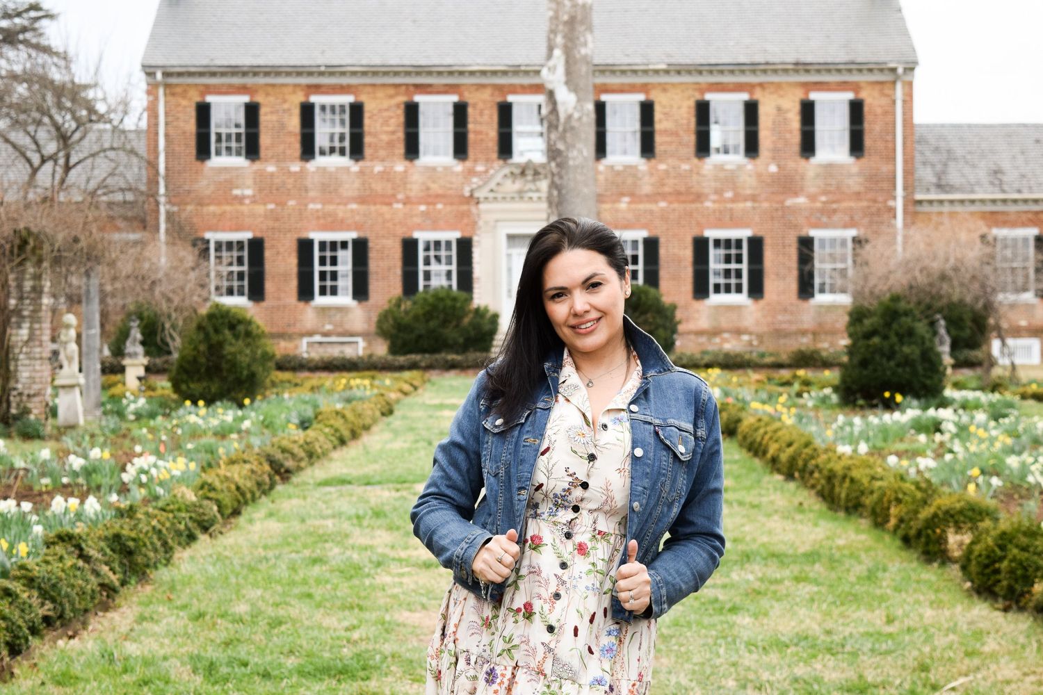 Woman in floral dress and denim jacket poses in formal garden before historic brick colonial mansion.