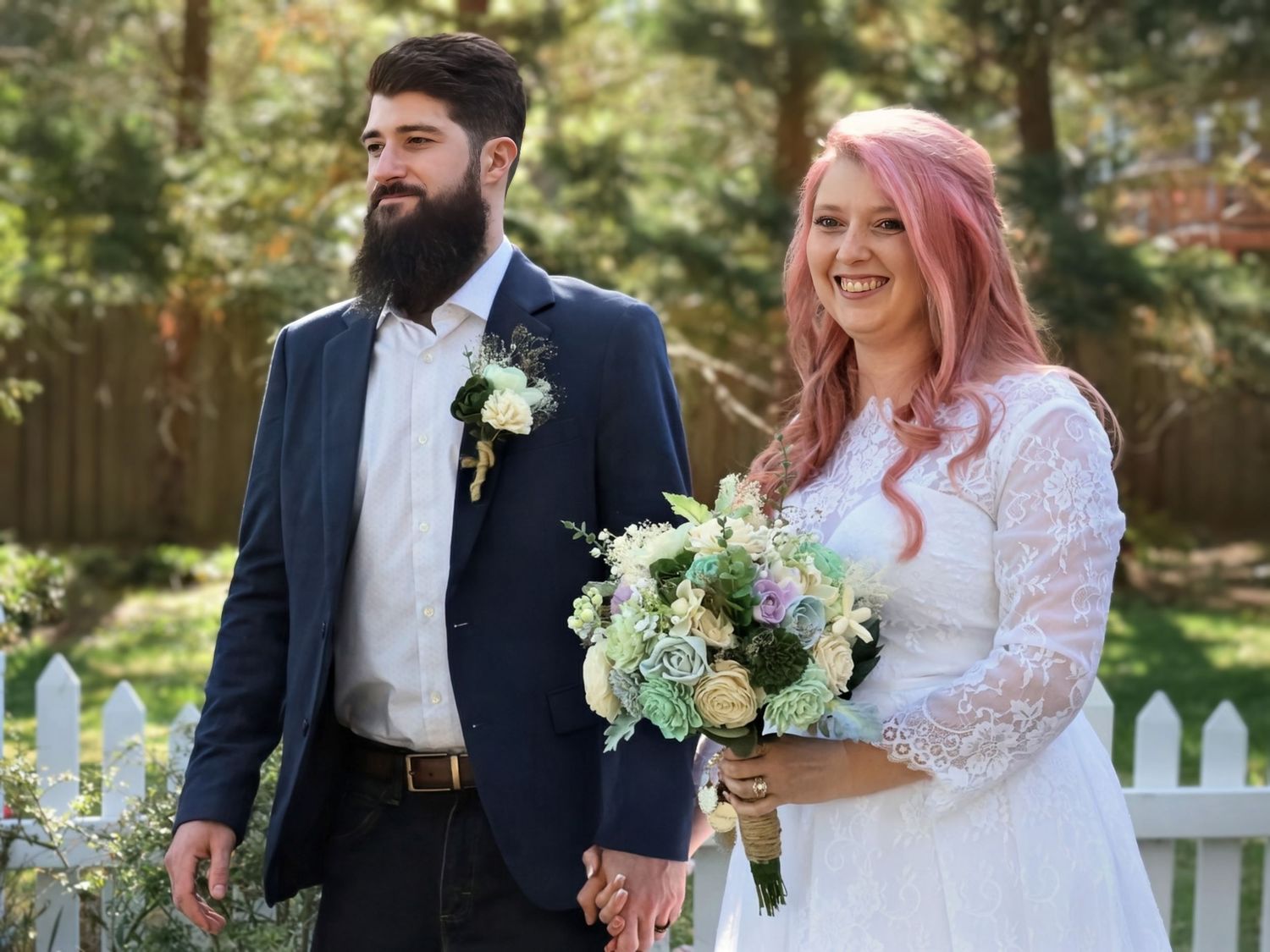 Bearded groom in navy suit walks with pink-haired bride in lace dress holding bouquet after outdoor ceremony.