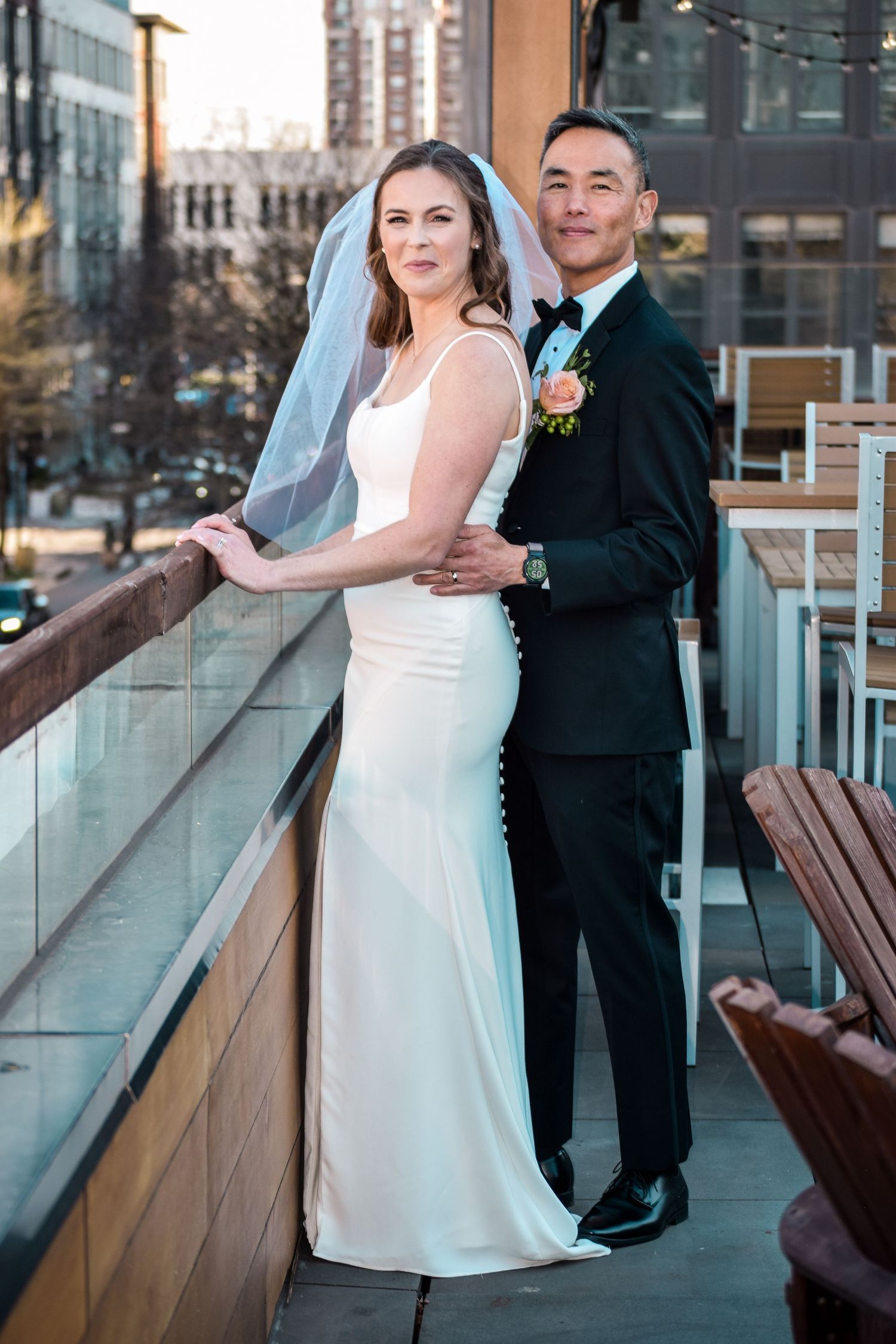 Bride and groom in elegant wedding attire pose on rooftop balcony with city skyline at dusk.