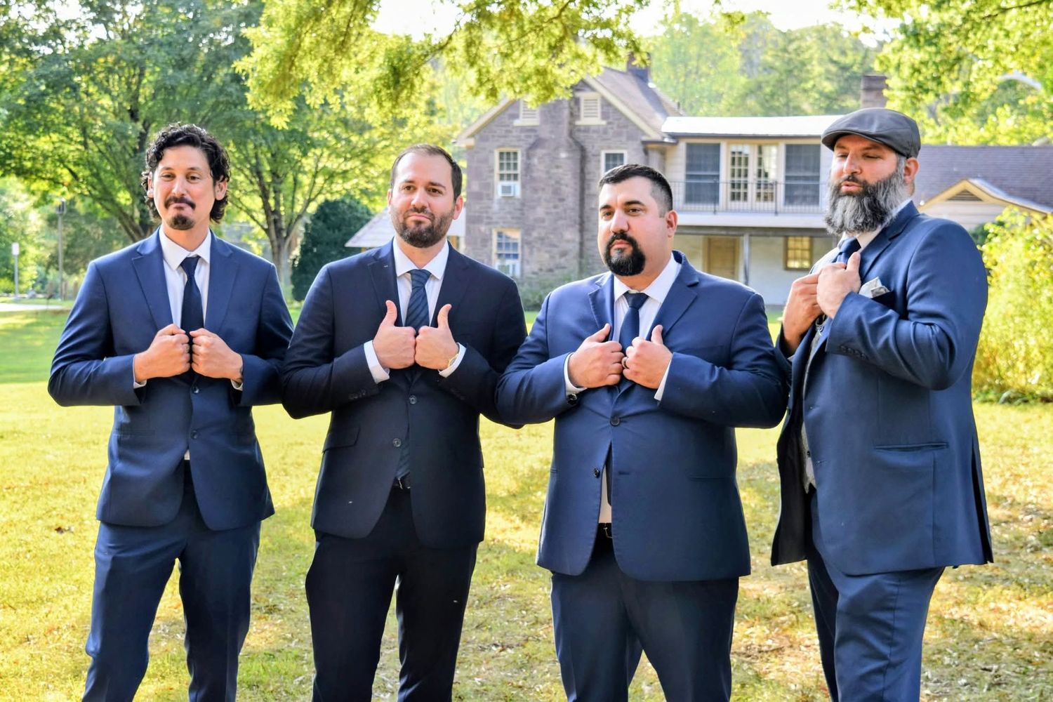 Four groomsmen in navy suits pose playfully with thumbs up outdoors on wedding day.