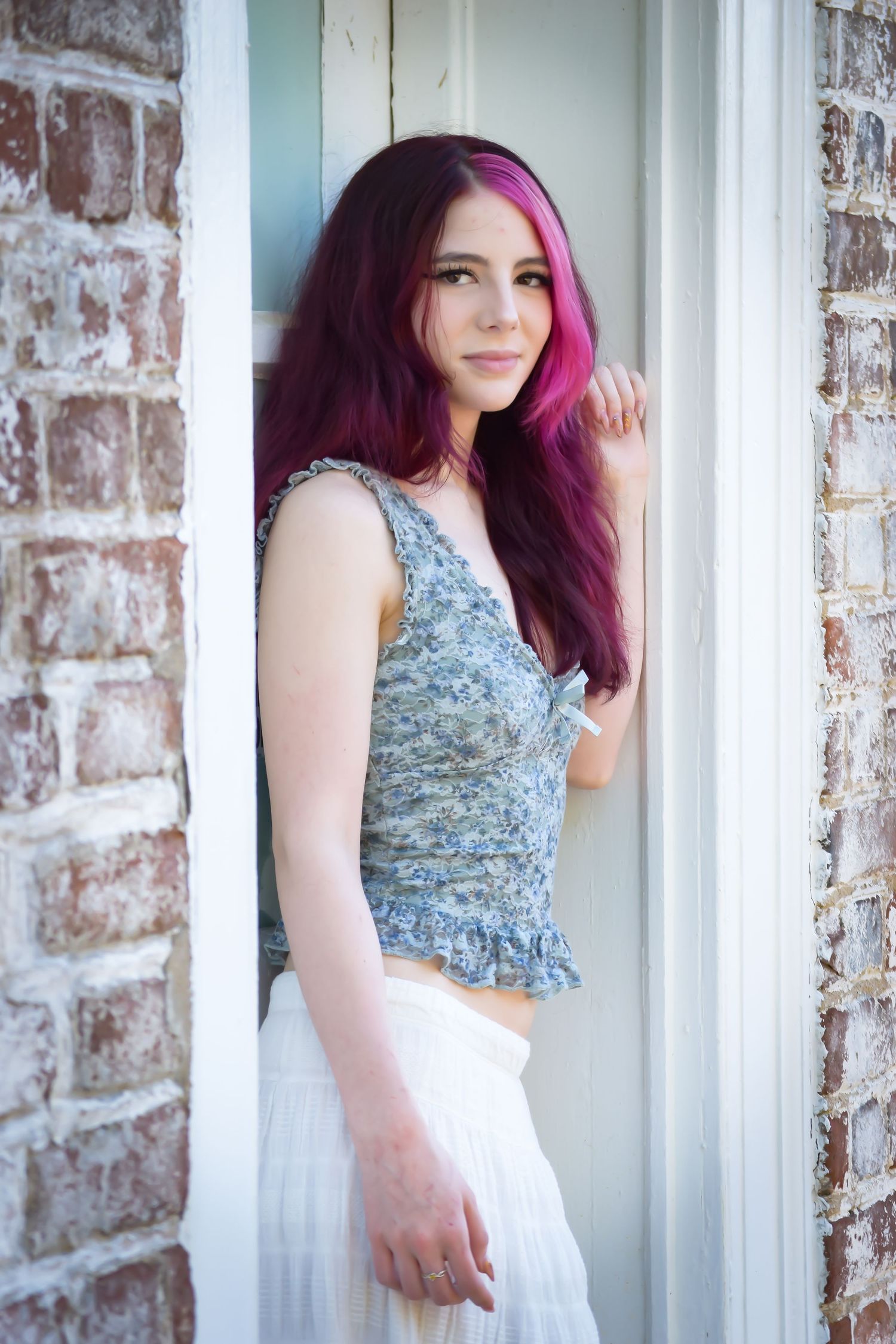 Young woman with vibrant pink-purple hair poses in a doorway wearing a floral crop top and white skirt.