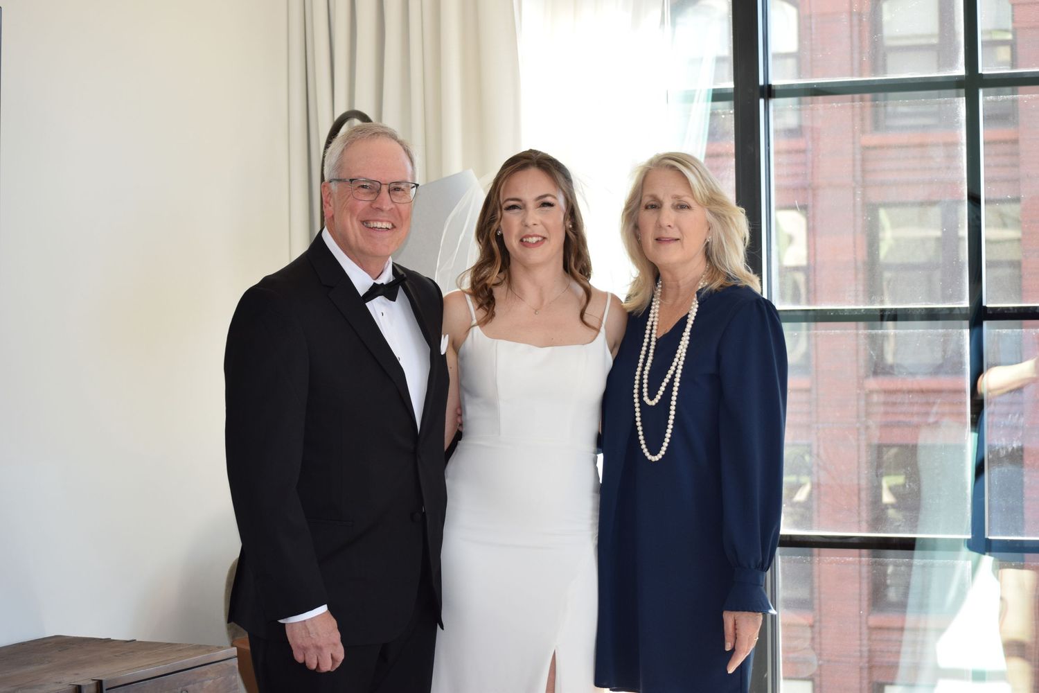Bride in white gown poses with parents, father in tuxedo and mother in navy dress with pearls.