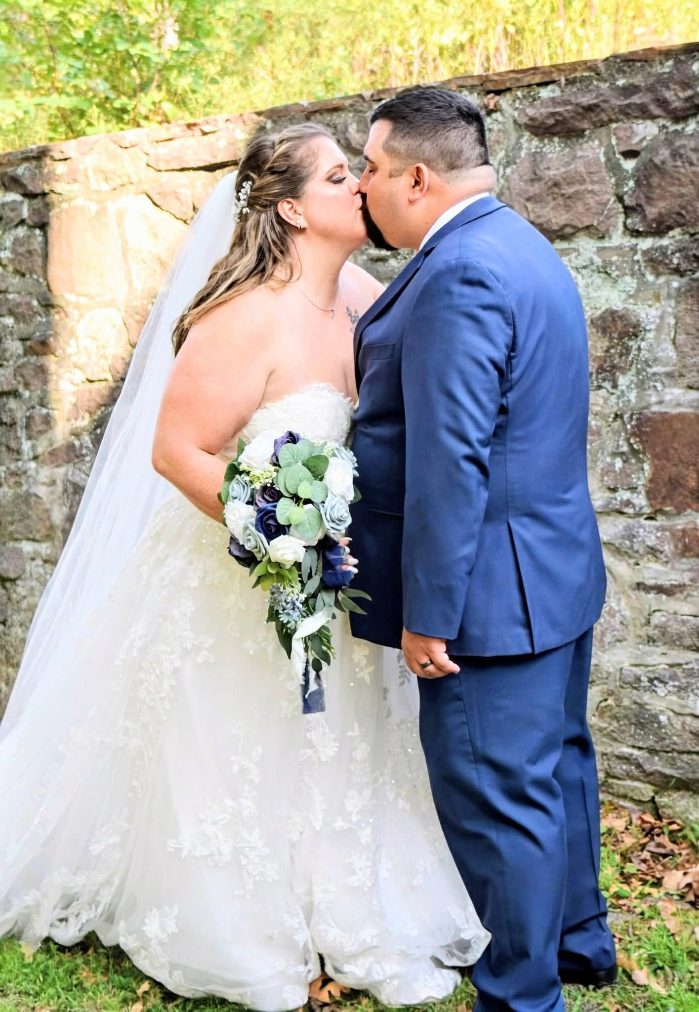 Bride and groom share a kiss in front of a stone wall, bride holding a white and blue bouquet.