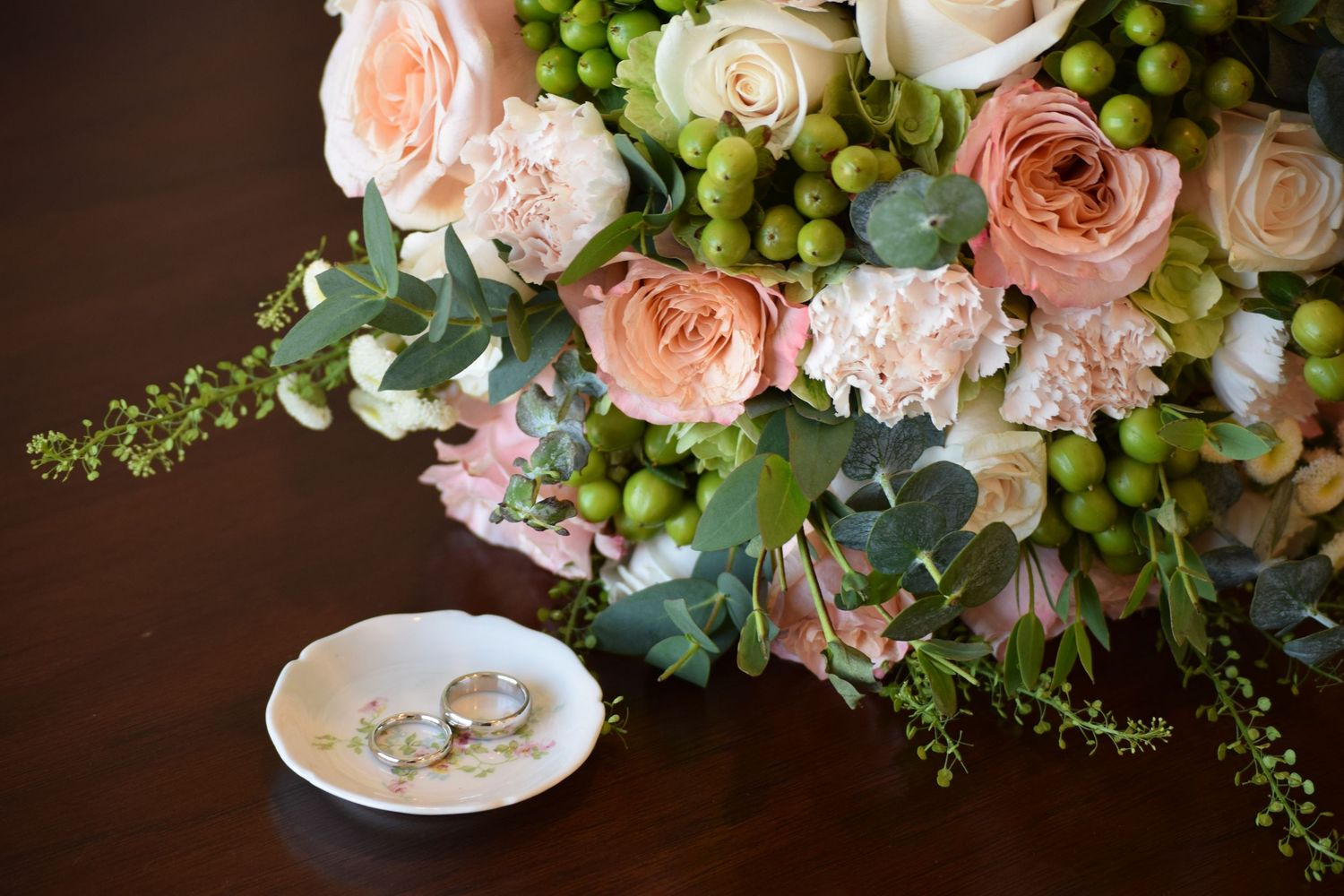 Elegant wedding bouquet with peach roses and greenery beside two gold rings.