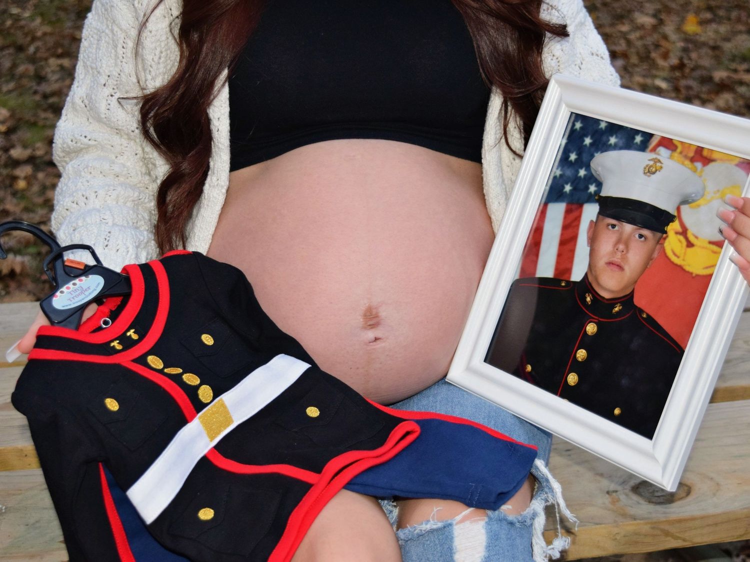 Pregnant woman holding a framed Marine portrait beside a dress uniform for a military maternity shoot.