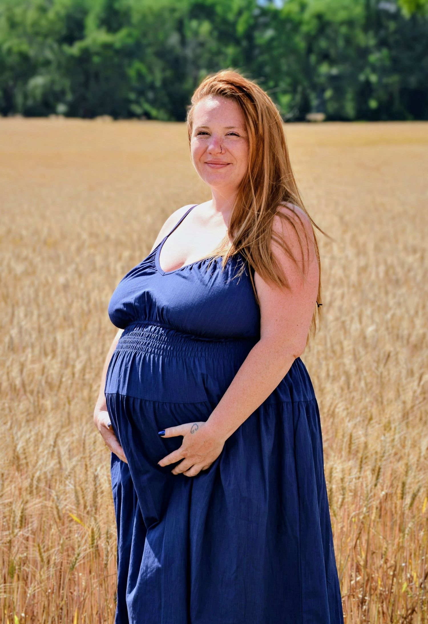 Pregnant woman in blue dress standing in golden wheat field, smiling outdoors on a sunny day.
