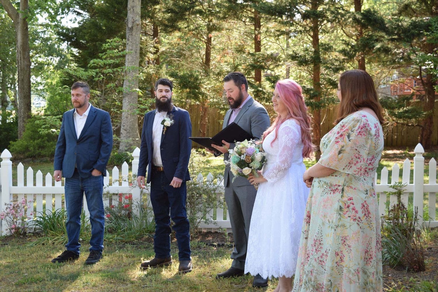 Small intimate outdoor wedding ceremony with couple exchanging vows before an officiant near a white picket fence.