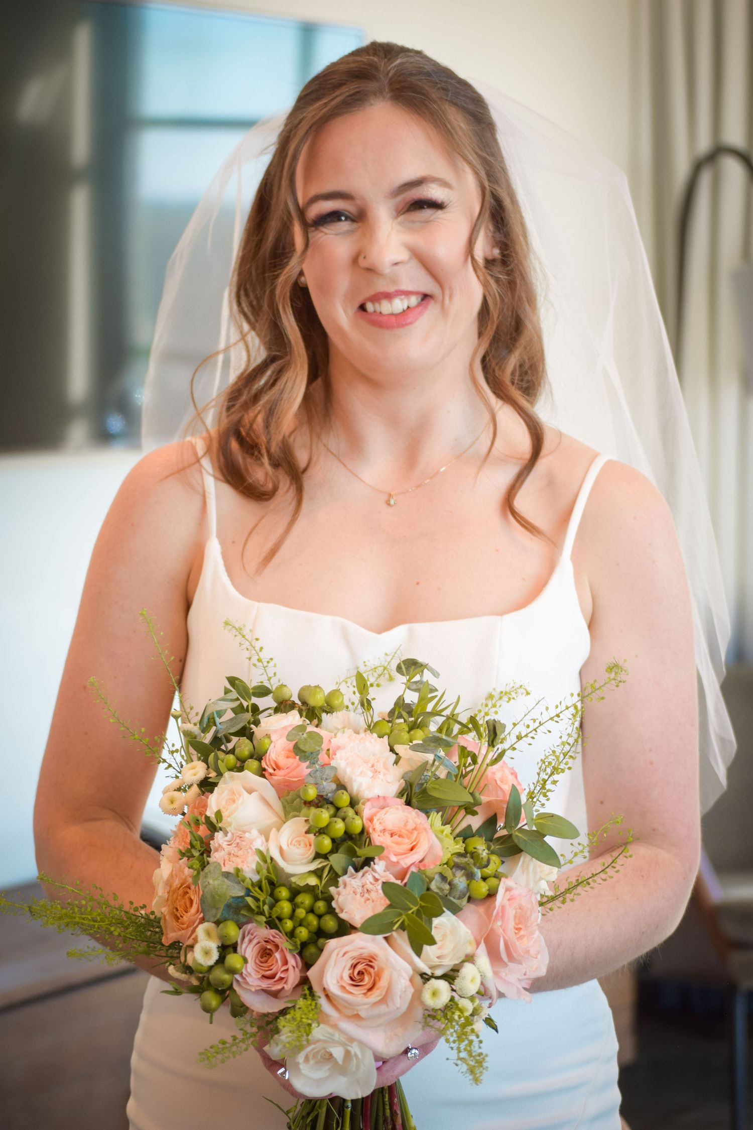 Smiling bride in white veil holding peach rose bouquet on her wedding day.