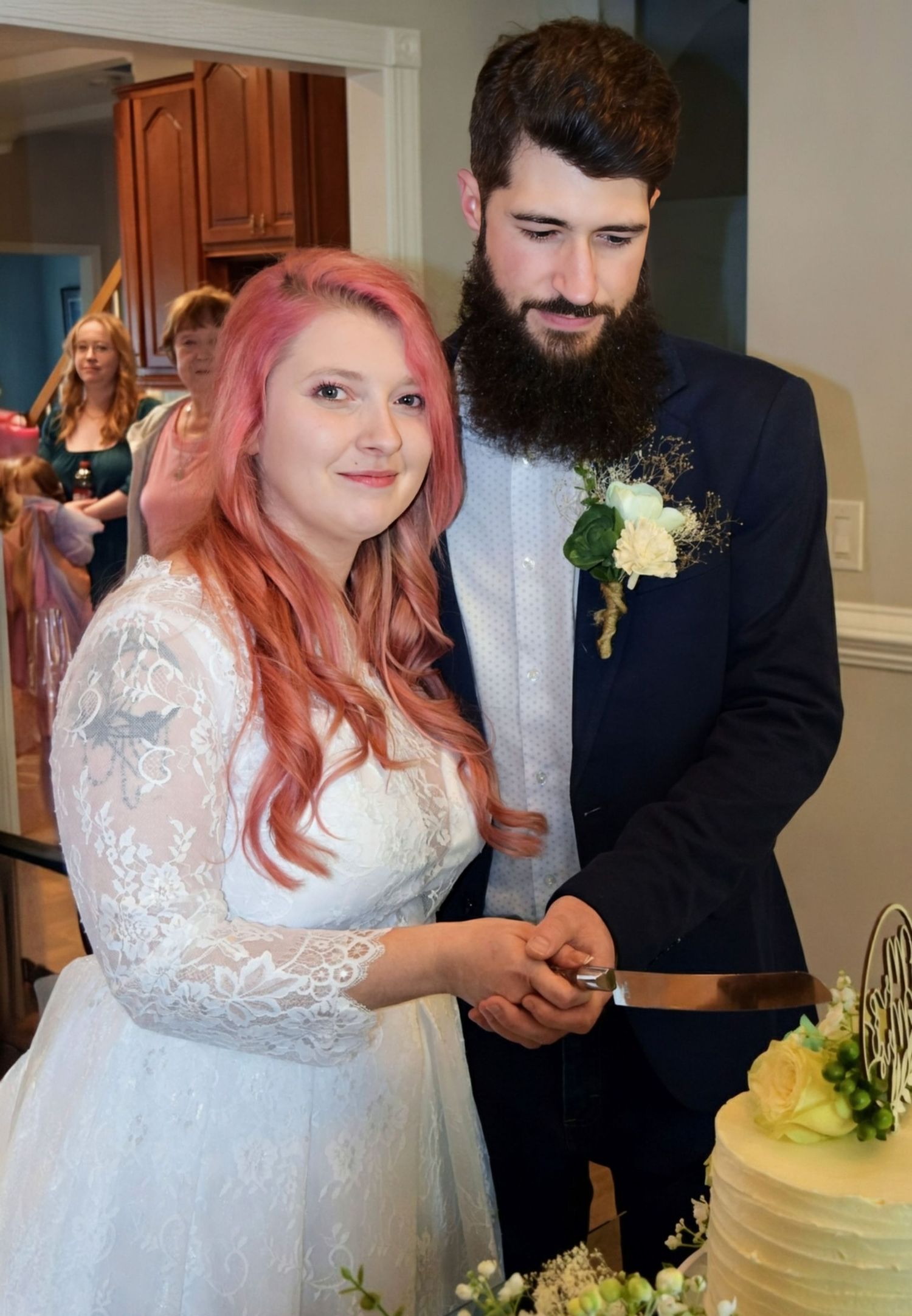 Newlywed couple cutting their white wedding cake together, pink-haired bride in lace dress and bearded groom in navy suit.