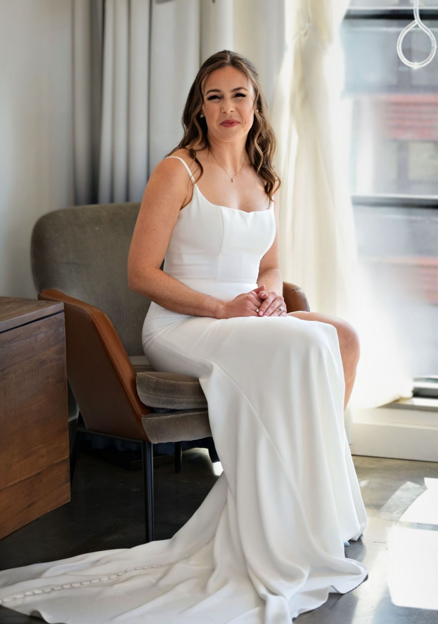 Elegant bride in a sleek white spaghetti-strap wedding gown sits gracefully in a chair by a window.