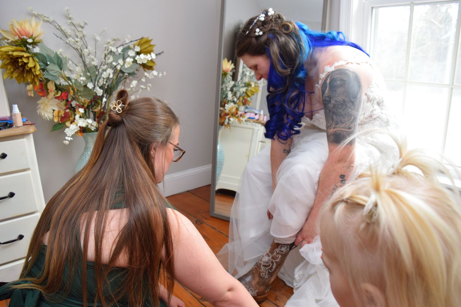 Bridesmaids kneeling to help tattooed bride with blue hair prepare her wedding dress.
