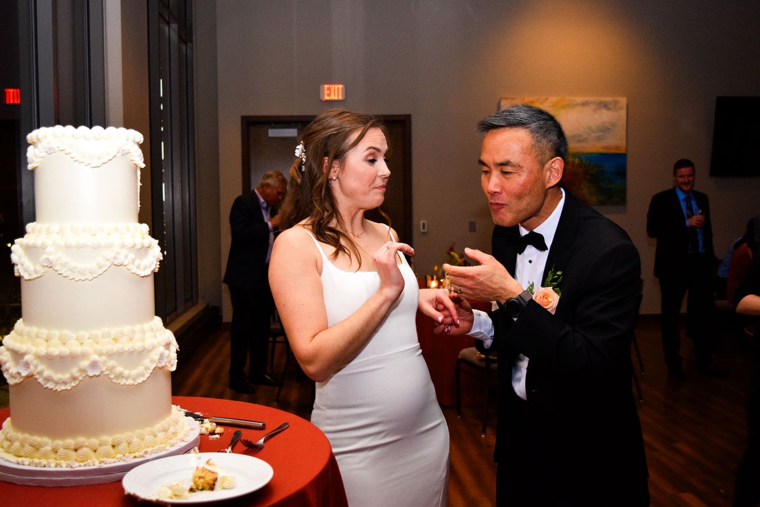 Newlyweds sharing wedding cake at reception beside elegant white tiered cake.