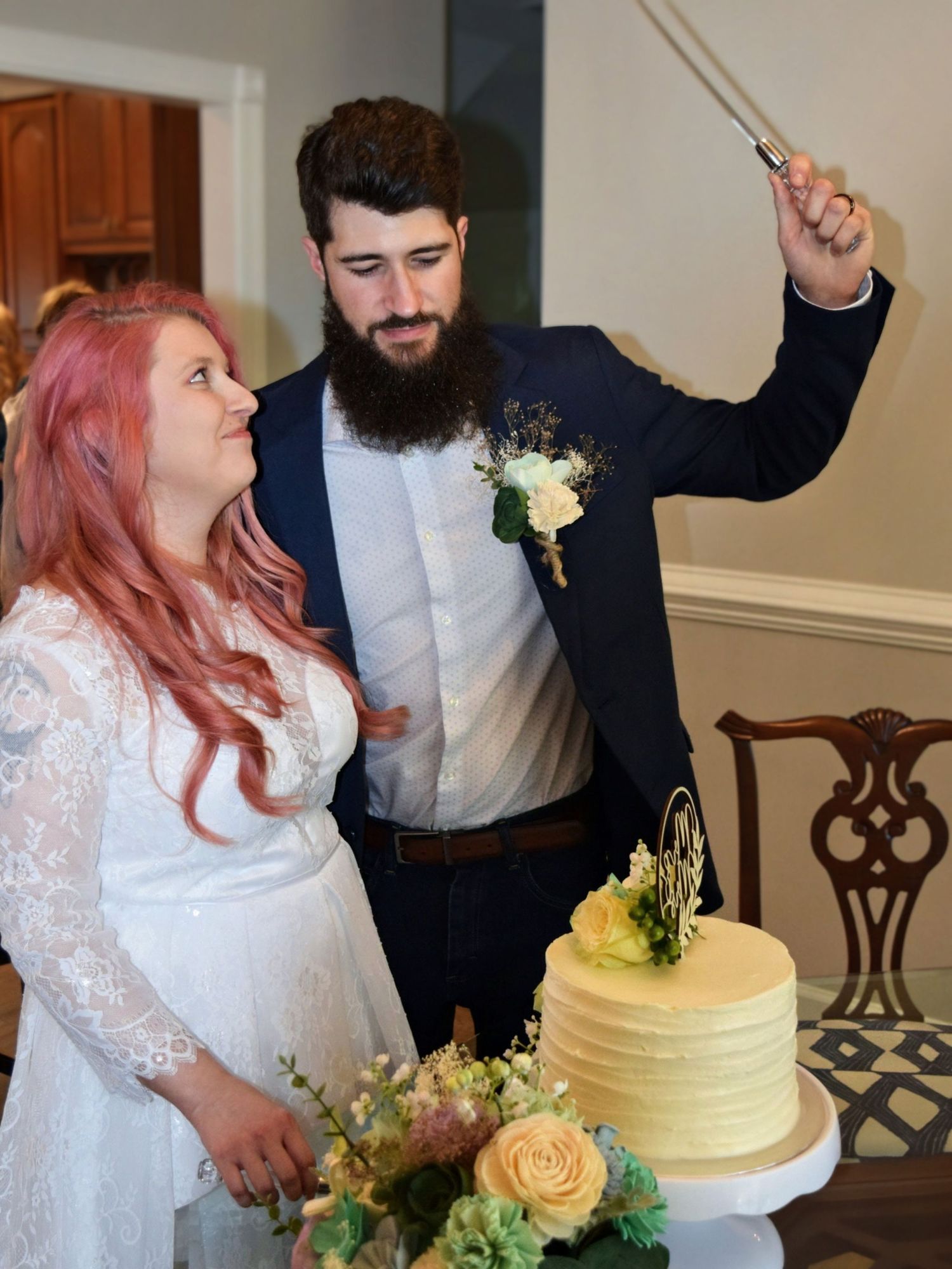 Bearded groom raising cake knife while pink-haired bride smiles beside yellow wedding cake at indoor reception.