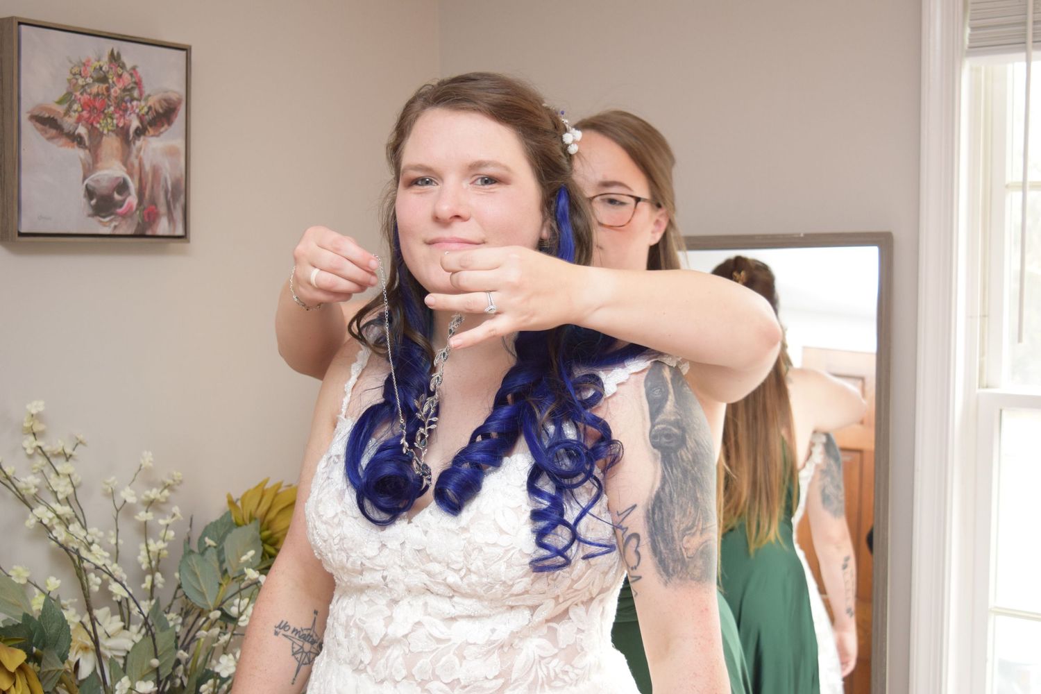 Bridesmaid helping blue-haired tattooed bride style her hair before wedding ceremony.