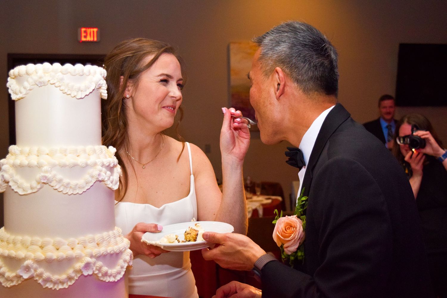 Bride feeding wedding cake to groom at elegant reception with white tiered cake.