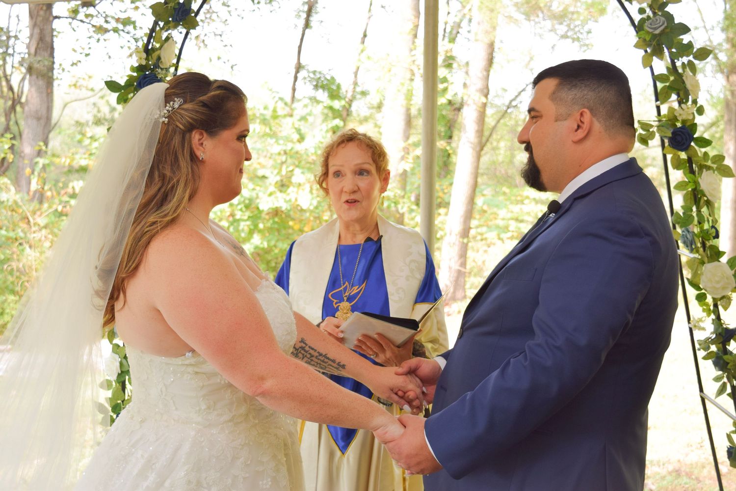 Bride and groom holding hands at outdoor wedding ceremony with officiant under a floral arch in the woods.
