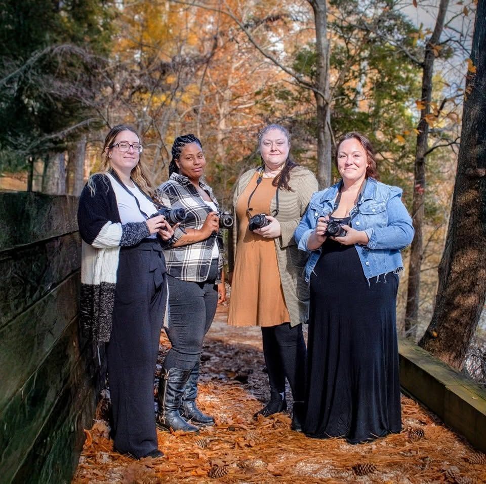 Four women photographers holding cameras pose together on a leaf-covered path surrounded by autumn foliage.