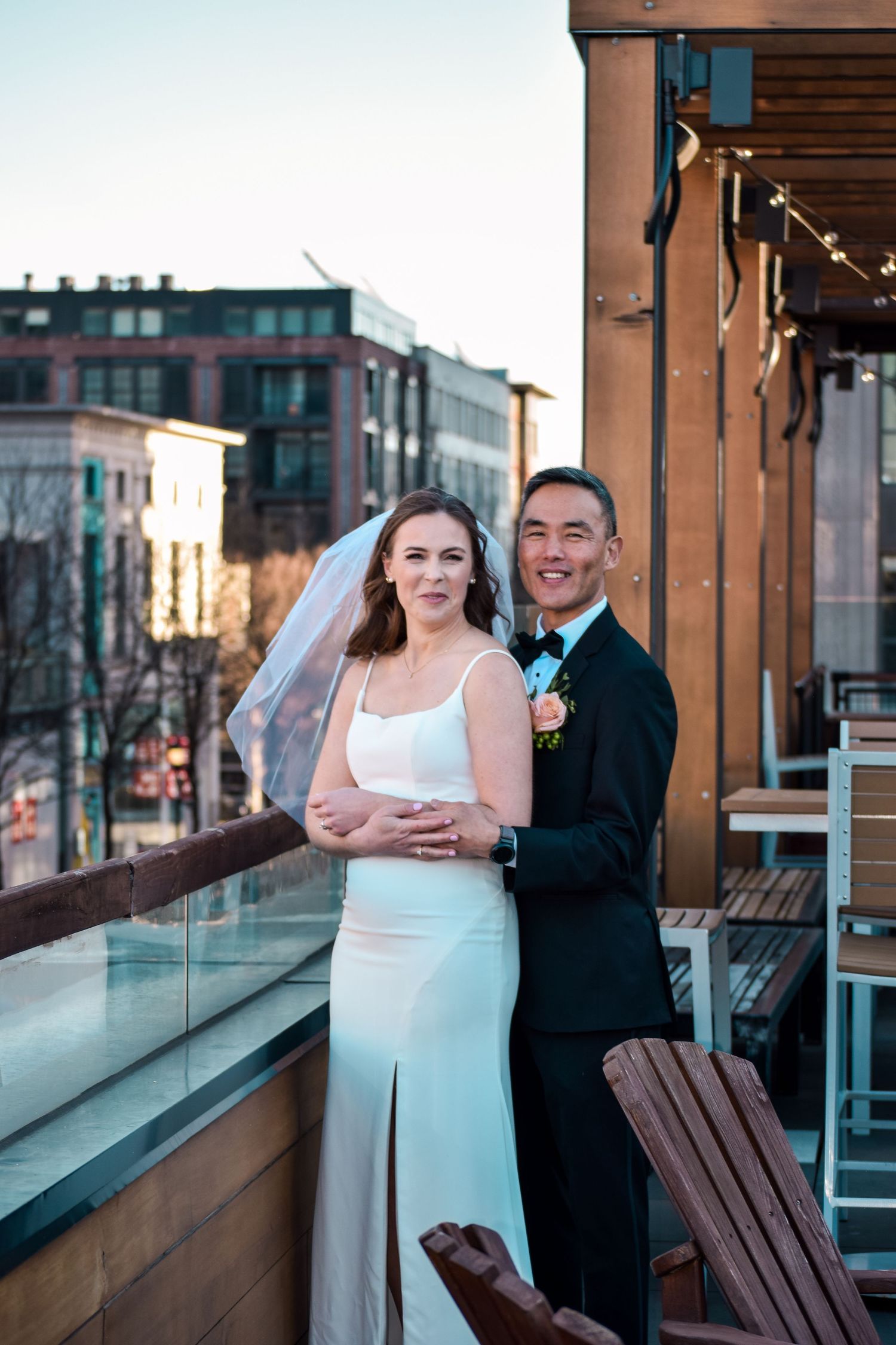 Newlyweds embrace on rooftop terrace at sunset with urban cityscape in the background.