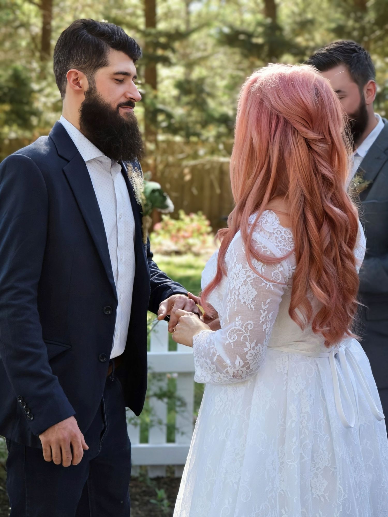 Bearded groom smiles as pink-haired bride places ring on his finger during outdoor backyard wedding ceremony.