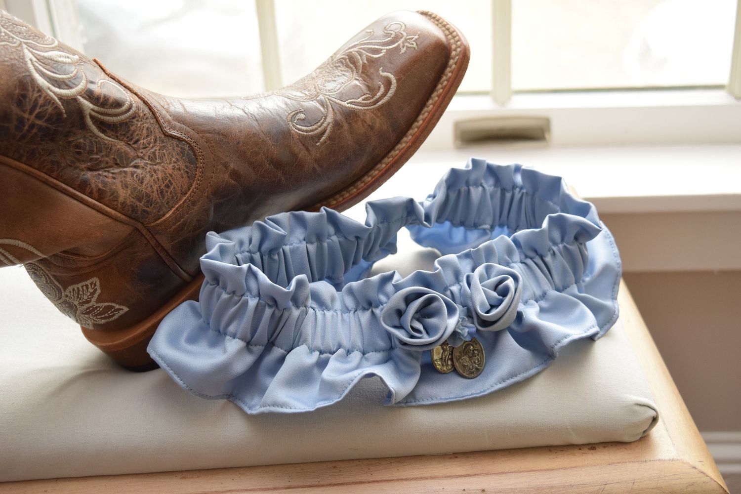 Blue satin wedding garter with roses and charm displayed next to brown cowboy boot.
