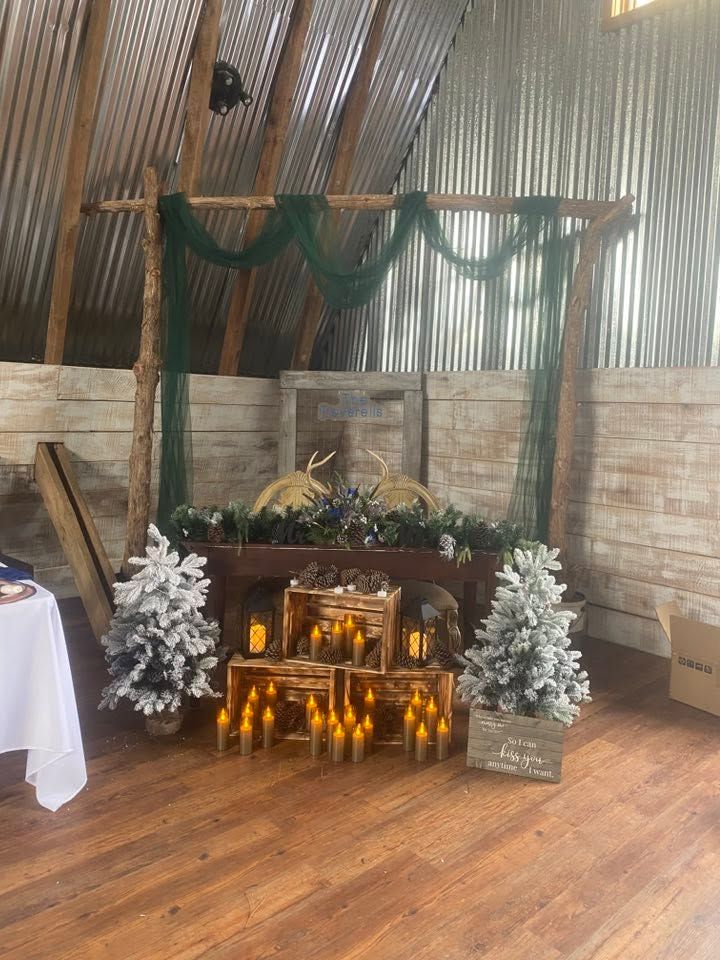 Rustic barn wedding backdrop with wooden arch, green draping, candles, and flocked Christmas trees.
