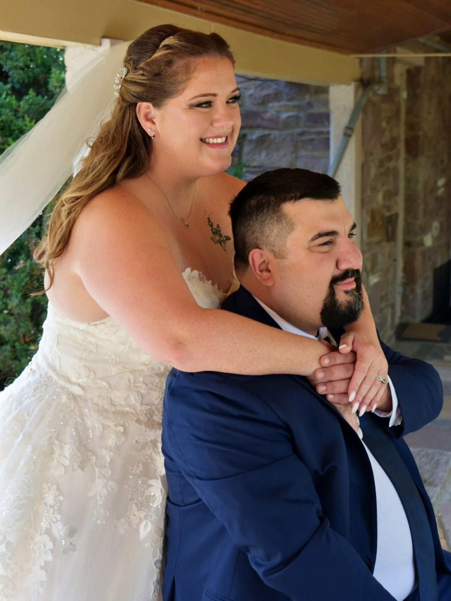 Bride in lace wedding gown embracing groom in navy suit under outdoor pavilion.