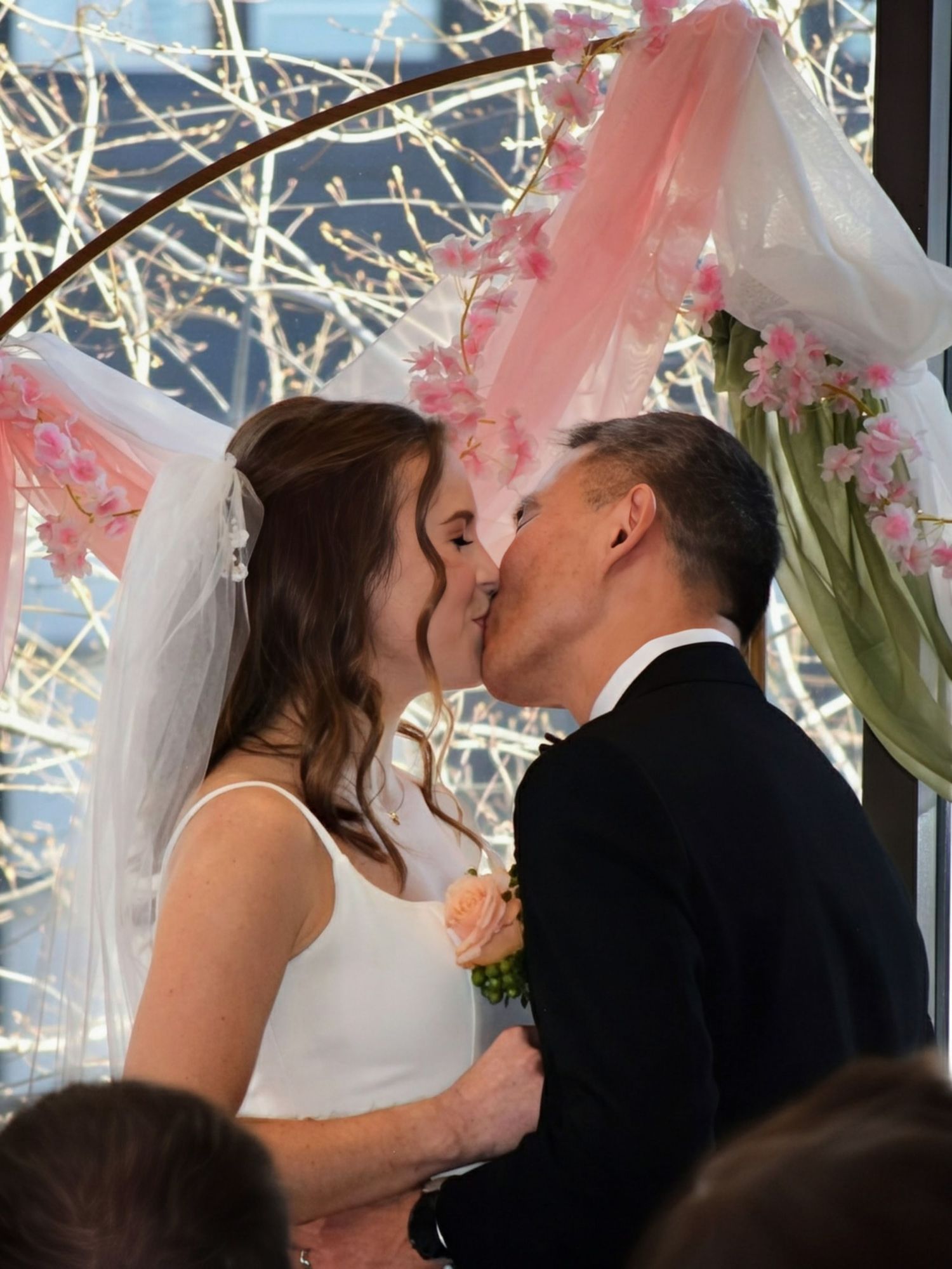 Newlyweds share romantic first kiss under decorative pink floral arch at wedding ceremony.