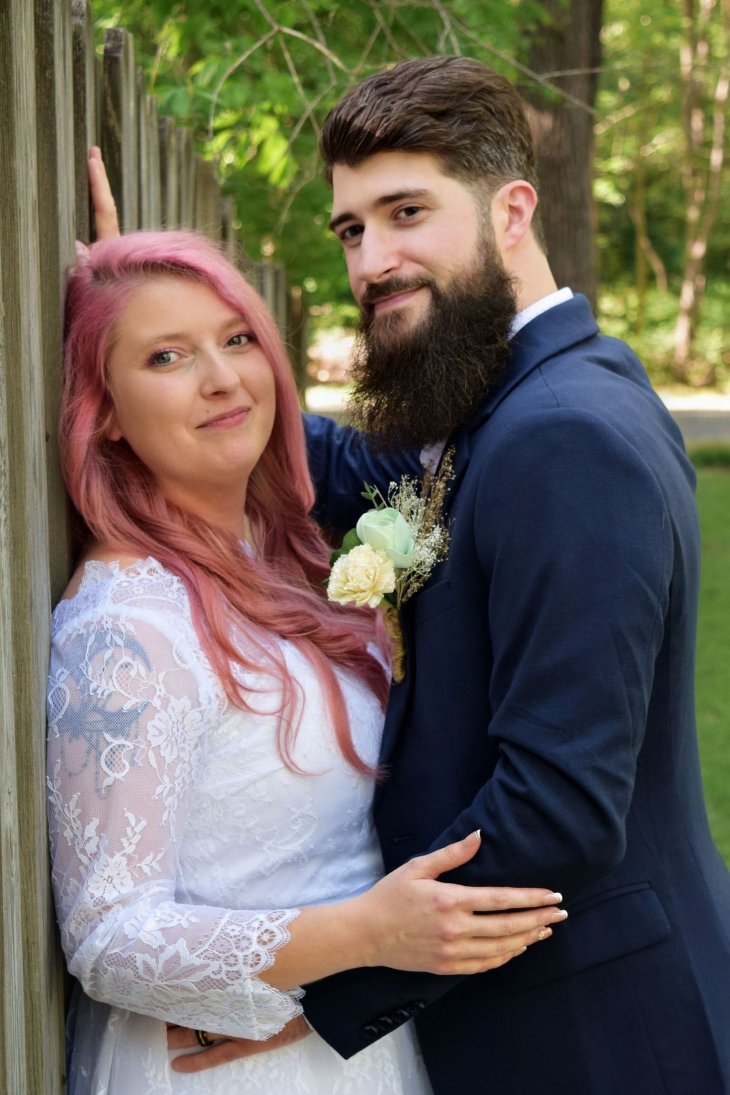 Bride with pink hair in lace dress and bearded groom in navy suit posing together against a wooden fence outdoors.