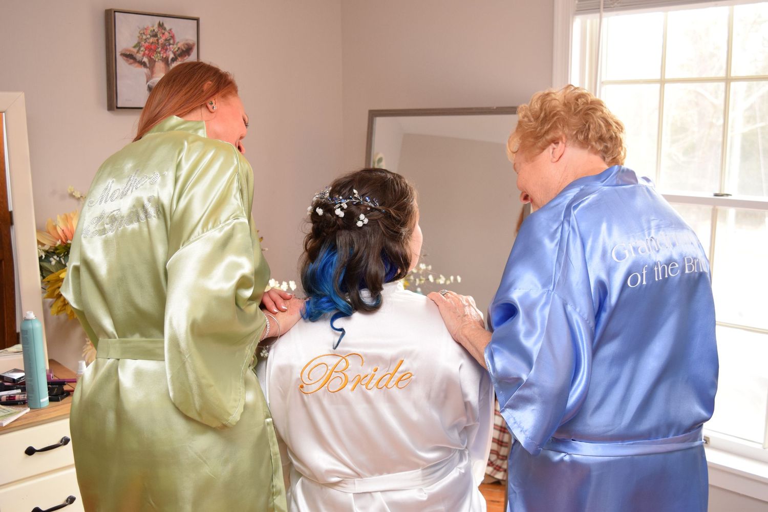 Bride in white satin Bride robe with blue hair flanked by two bridesmaids in satin robes during preparations.