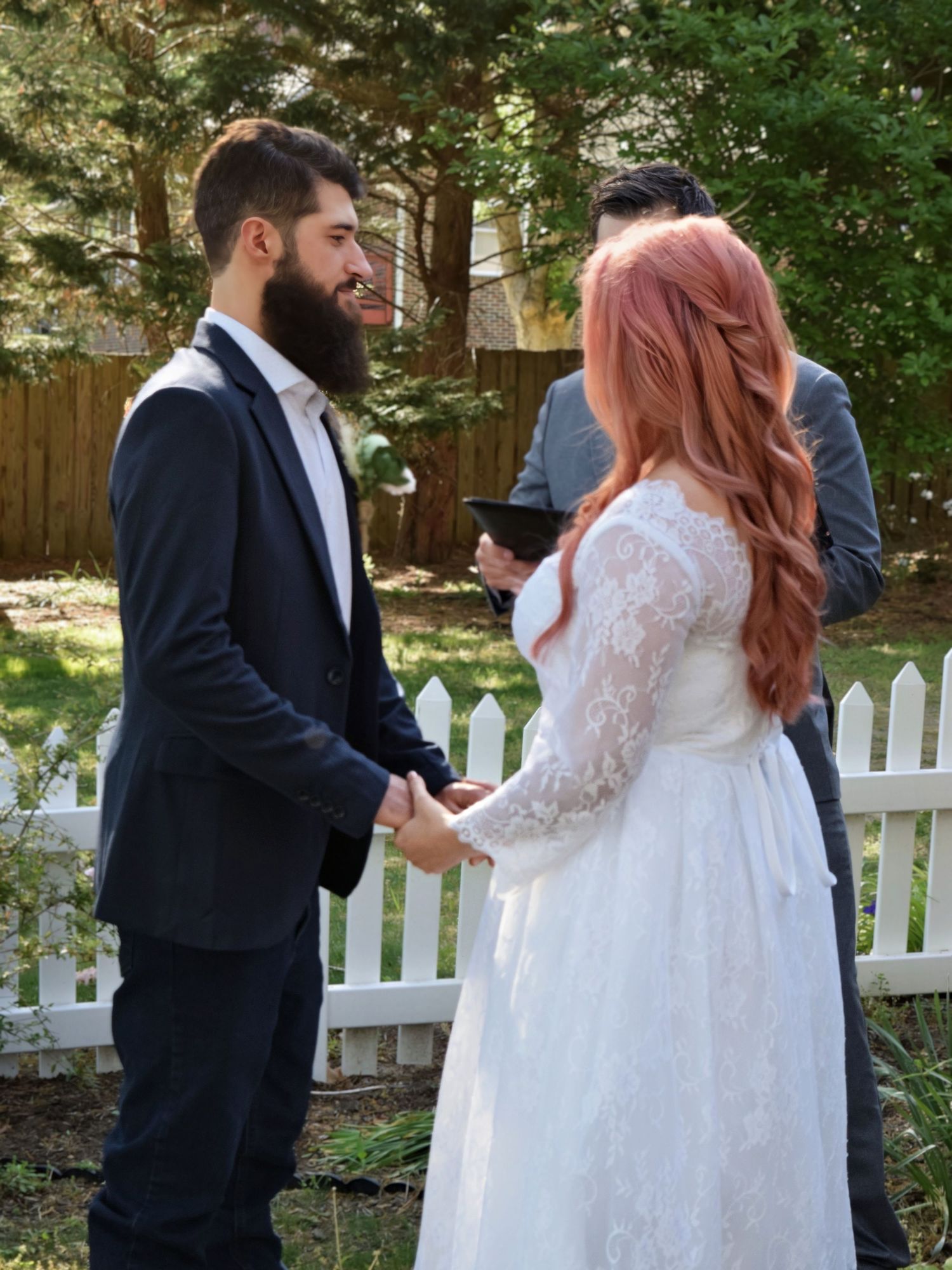Bearded groom holds hands with pink-haired bride during heartfelt outdoor wedding ceremony vows exchange.
