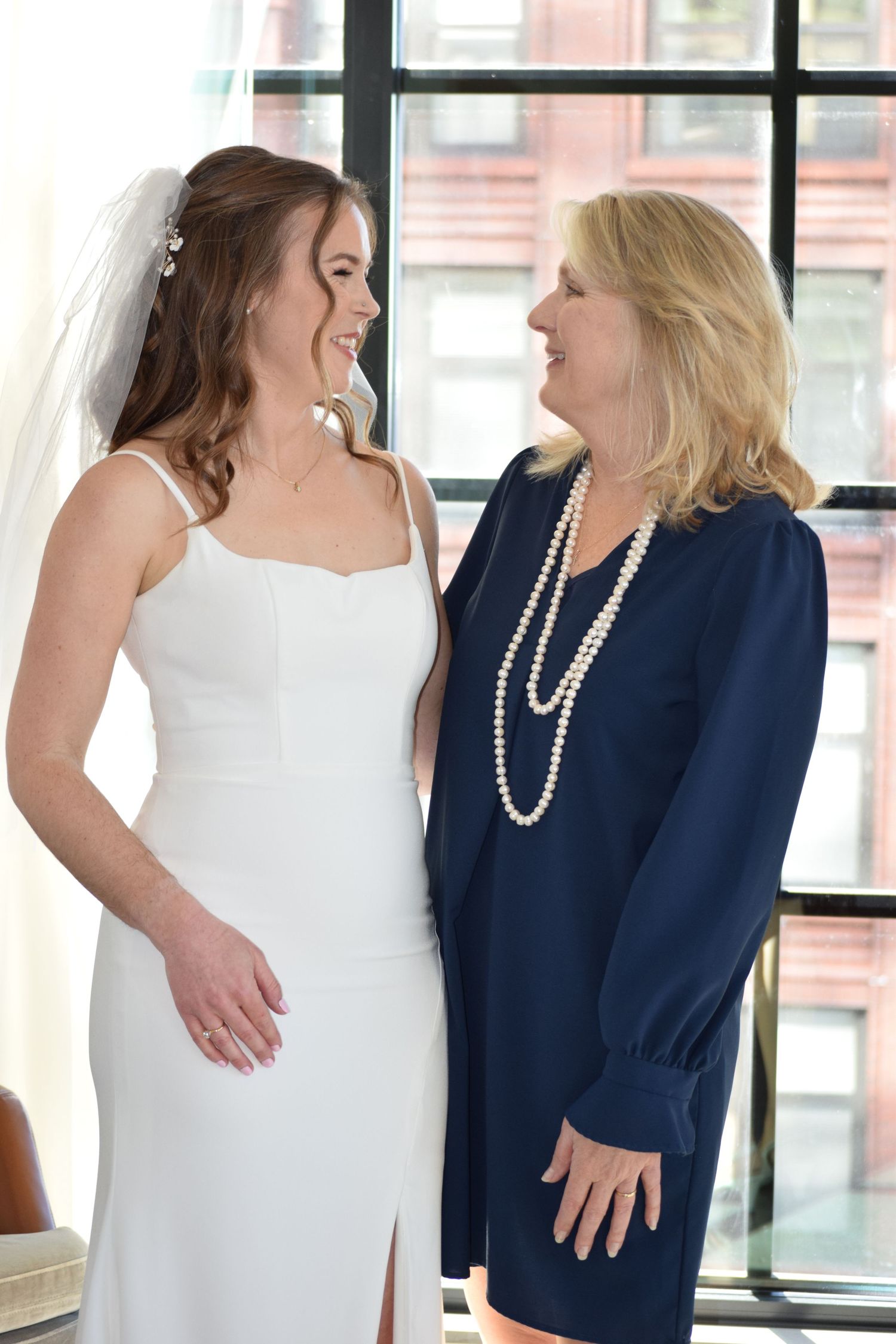 Bride in white gown and veil smiling with older woman in navy dress and pearl necklace at wedding.