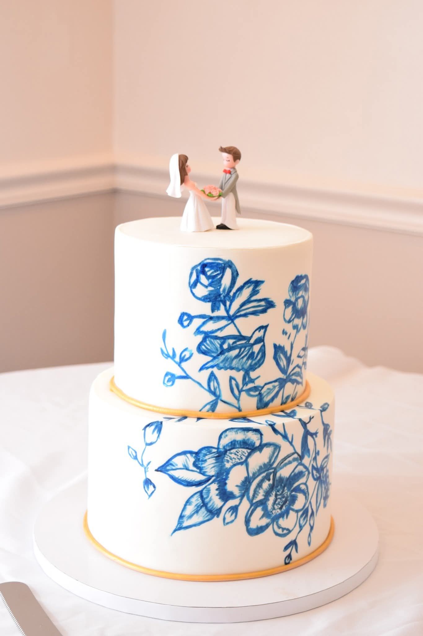 Two-tier white wedding cake with blue hand-painted floral designs and couple cake topper.